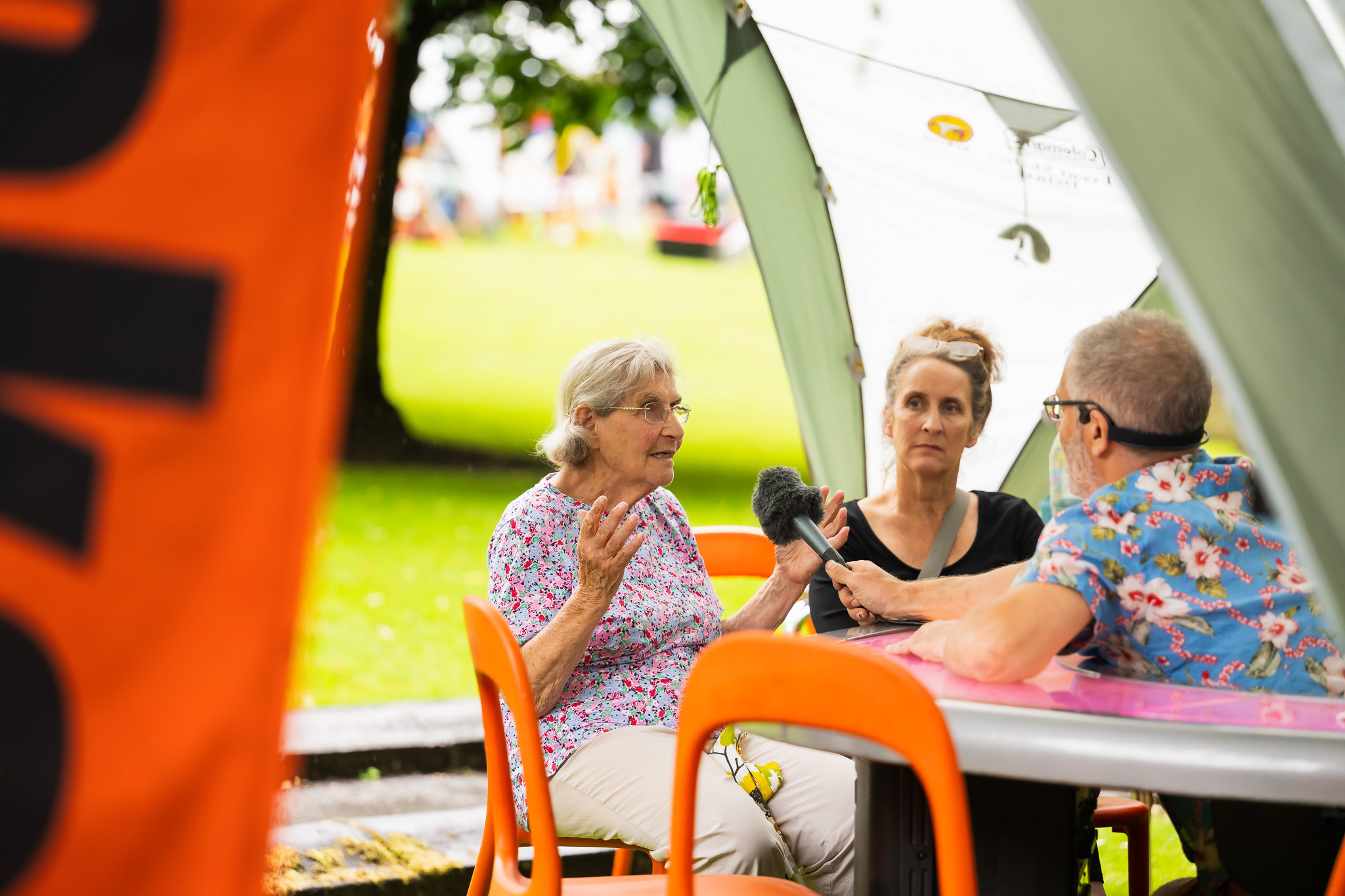 An older woman speaks whilst being interviewed by a man holding a microphone at an outdoor event; another woman listens nearby under a marquee, with orange chairs and a park in the background.