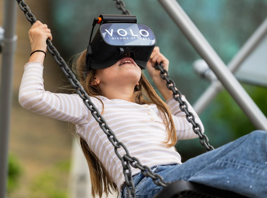 A young girl wearing a VR headset labelled VOLO dreams of flight sits on a swing, holding the chains and leaning back, appearing to enjoy a virtual reality experience outdoors.