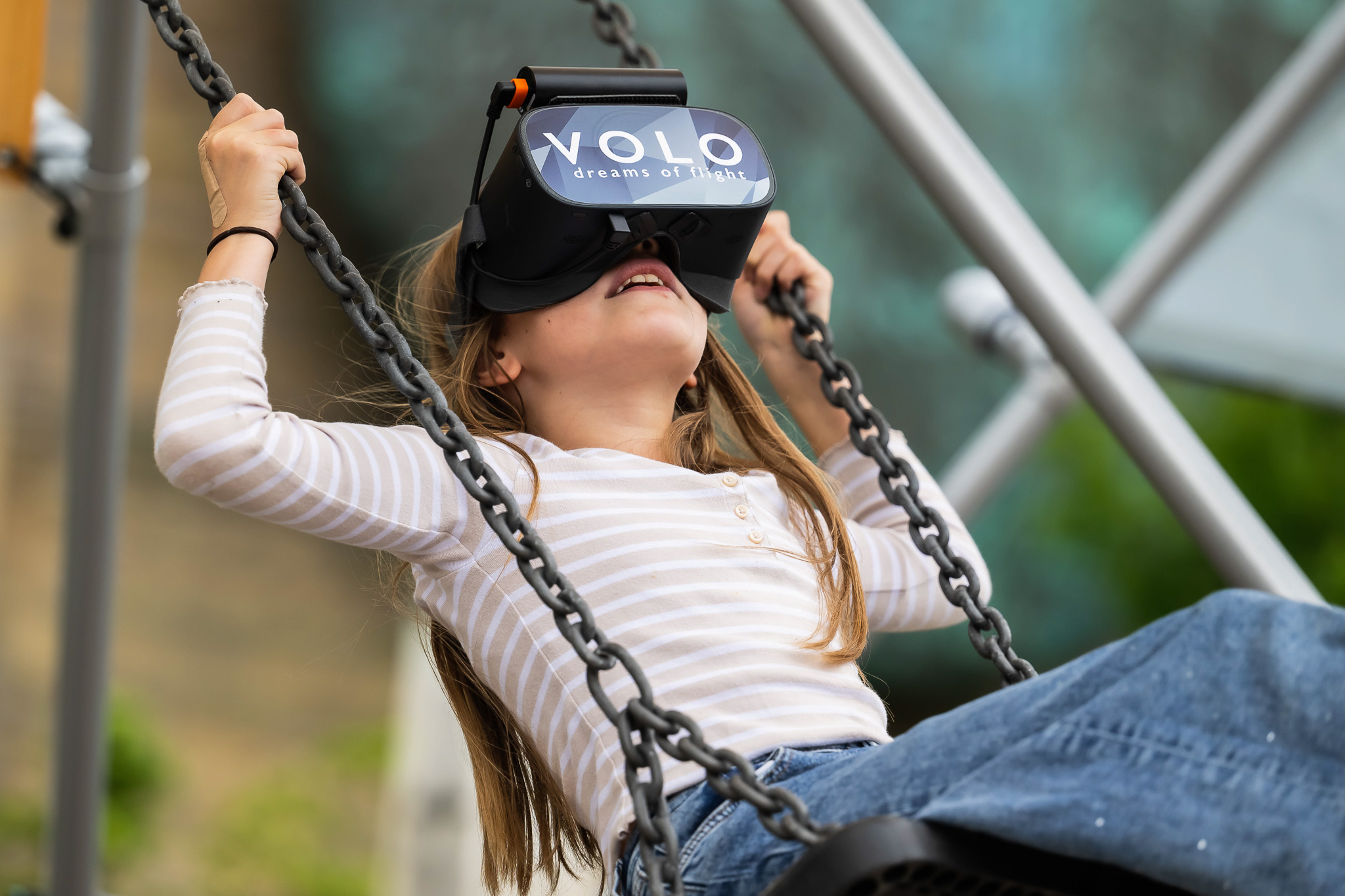 A young girl wearing a VR headset labelled VOLO dreams of flight sits on a swing, holding the chains and leaning back, appearing to enjoy a virtual reality experience outdoors.