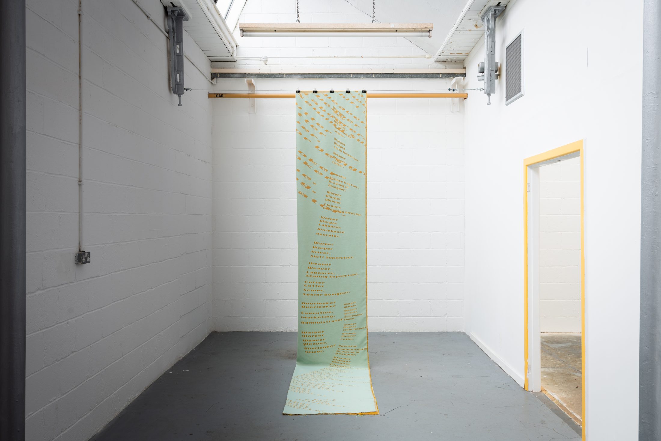 A long, vertical fabric banner with text hangs from a wooden rod in a white, minimalist room with high ceilings and grey floors. The banner extends from the rod to the floor, partially unrolled.