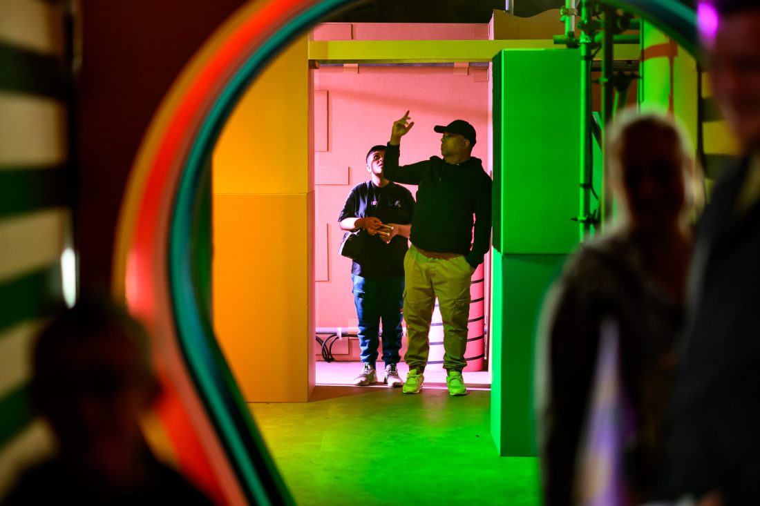 Two people stand in a brightly coloured, geometric corridor. One person points upwards while the other looks at him. Neon lights and rainbow arches frame the scene, with silhouettes of other people in the foreground.