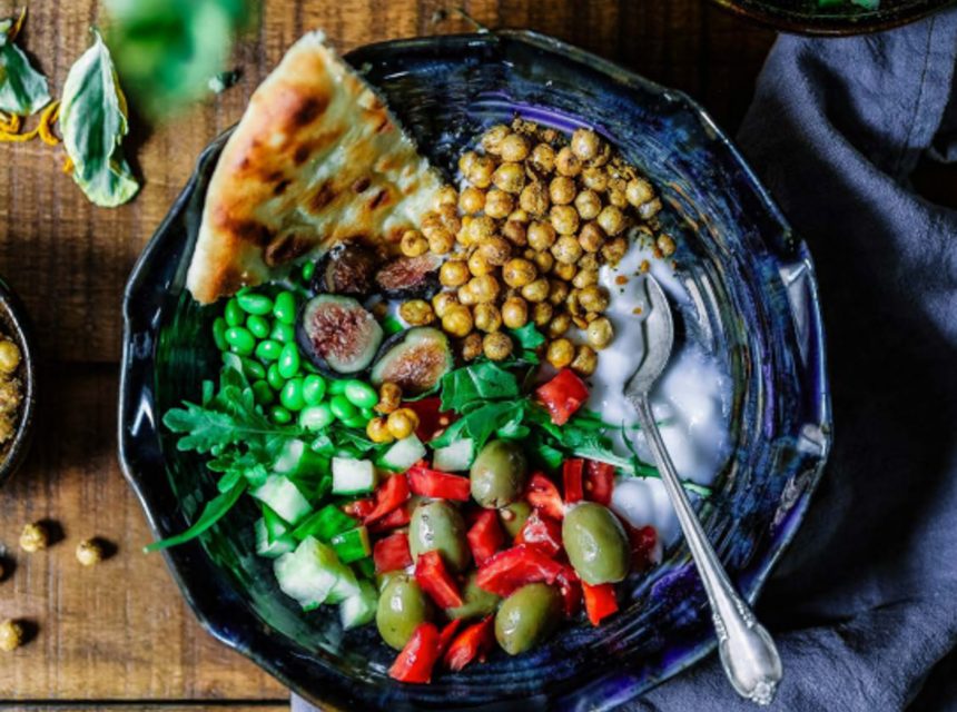 A colourful bowl with chickpeas, flatbread, peas, fresh greens, cucumber, red pepper, green olives, figs, and a creamy white sauce, served with a silver spoon on a dark ceramic plate.