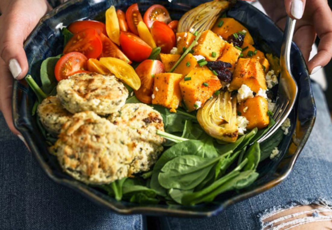 A person holds a bowl filled with fresh spinach, falafel patties, colourful cherry tomatoes, roasted butternut squash, onions, and crumbled feta cheese, with a fork resting inside.