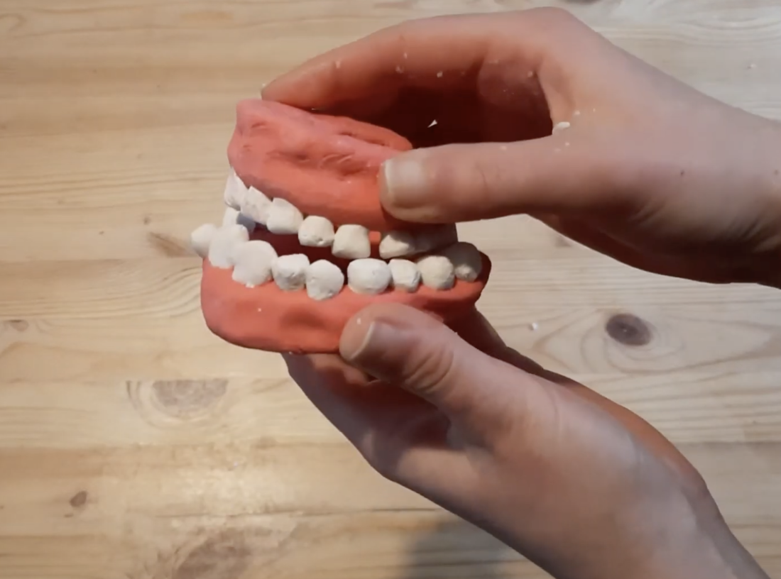 Two hands hold a model of human teeth and gums, made from a pink and white material, over a wooden surface. The model resembles a simplified version of a set of dentures.