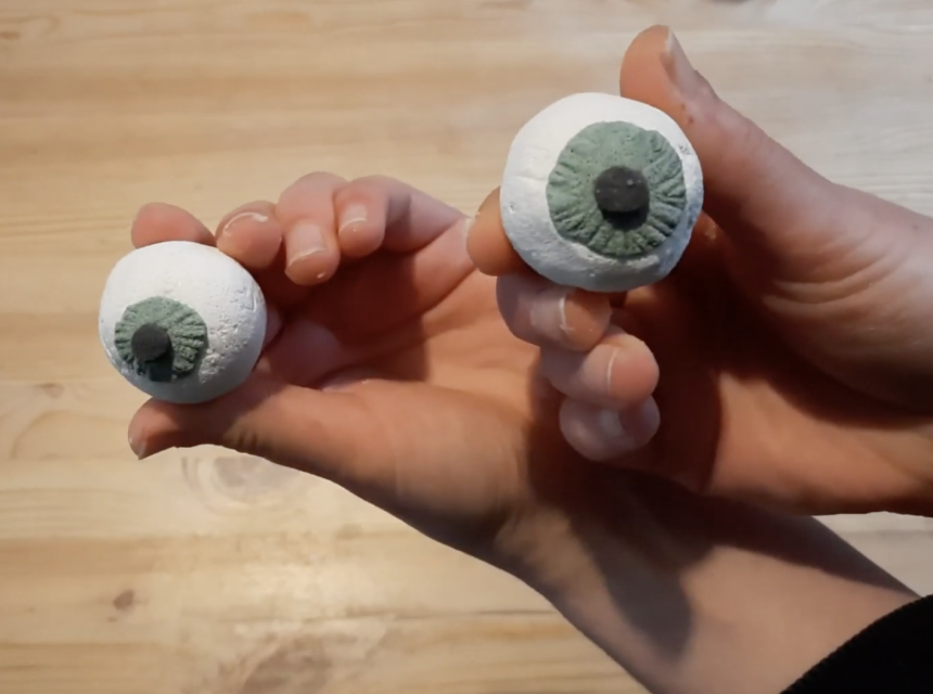 Two hands holding realistic clay eyeball models with green irises and black pupils over a light wooden surface.