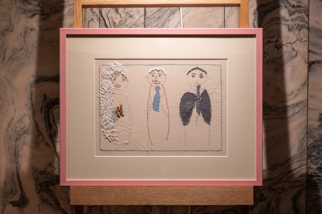 Framed artwork with a pink border depicts three simple, childlike figures; the left figure features textured white fabric, whilst the other two figures are drawn with coloured threads, all set against a beige background.