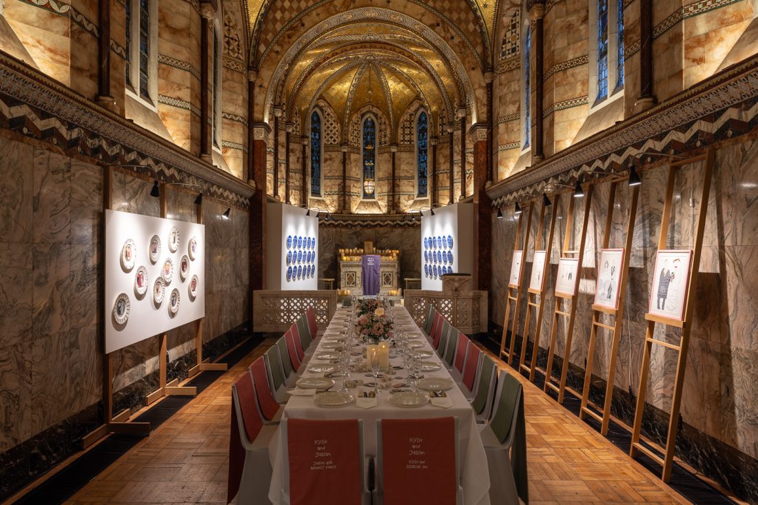 A long, elegant dining table is set with fine china and flowers inside a grand, ornately decorated hall with arched, gold-accented ceilings, marble walls, and framed artworks on easels along the sides.