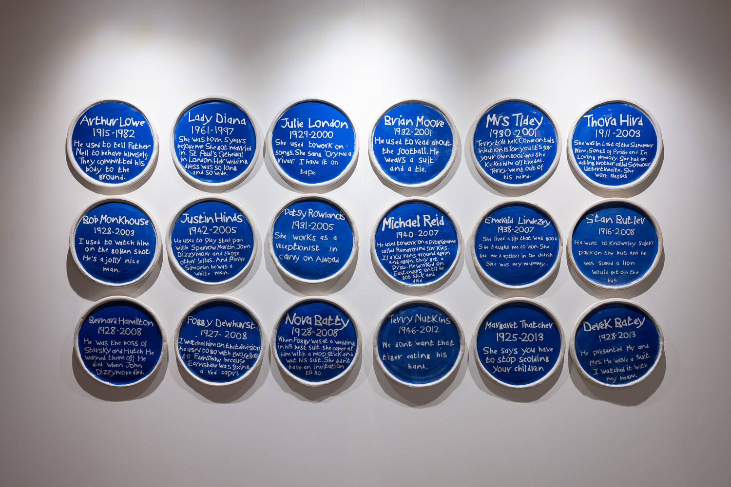 A wall display of sixteen blue circular plaques, each with handwritten white text commemorating different individuals and their life dates, mounted in four rows under spotlights.