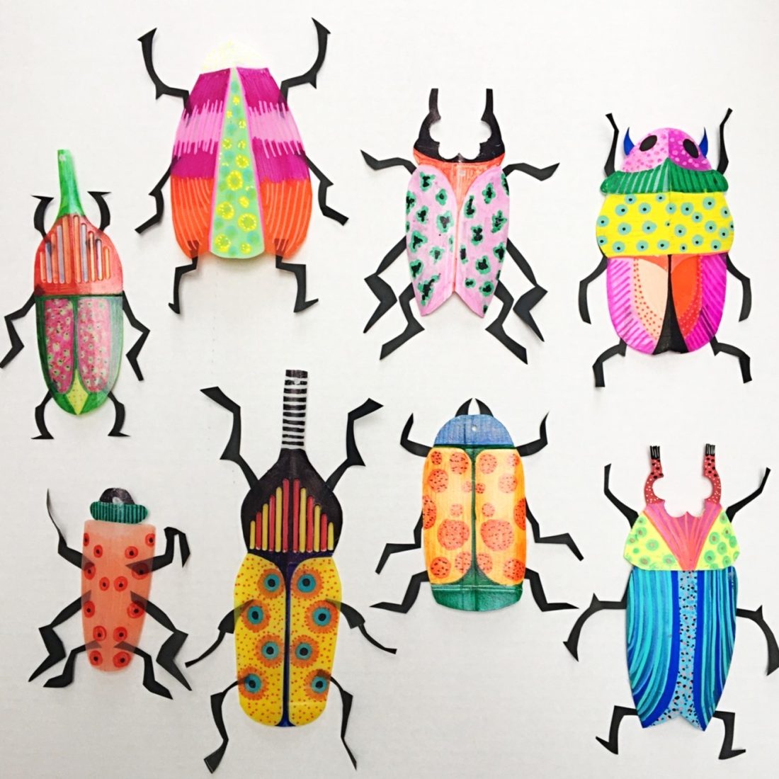Eight colourful, patterned paper cut-out beetles with black legs are arranged on a white background. Each beetle features unique shapes and bright designs, including stripes, dots, and geometric patterns.