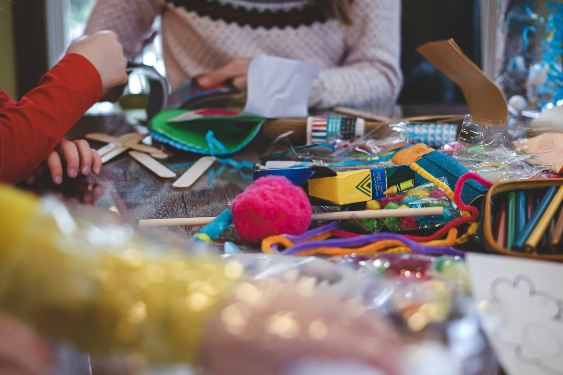 Children's hands work on a craft project at a table covered with colourful supplies, including crayons, lolly sticks, pipe cleaners, felt, and paper. An adult is partially visible in the background, supervising.