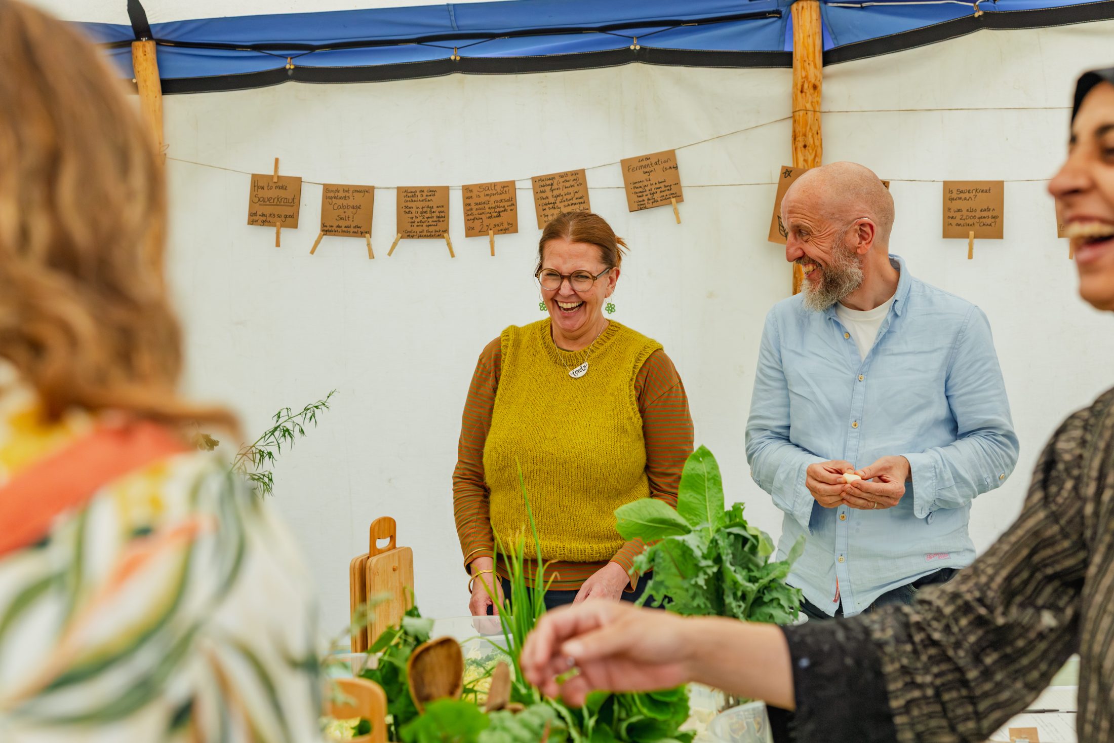 People laughing at a workshop with fresh greens and recipe cards hanging behind them.
