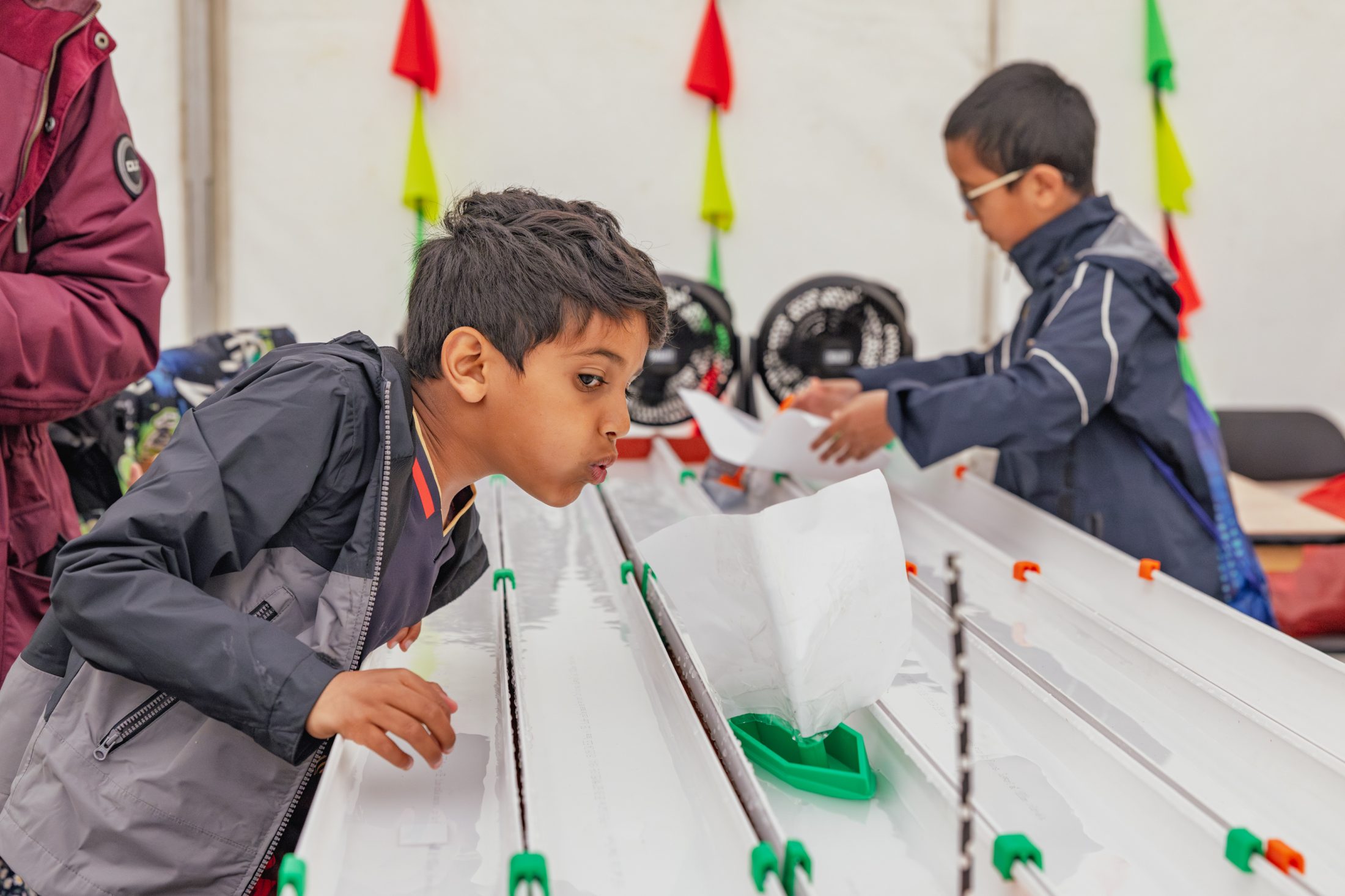 Two boys race paper sailboats on water tracks, using fans to propel them.