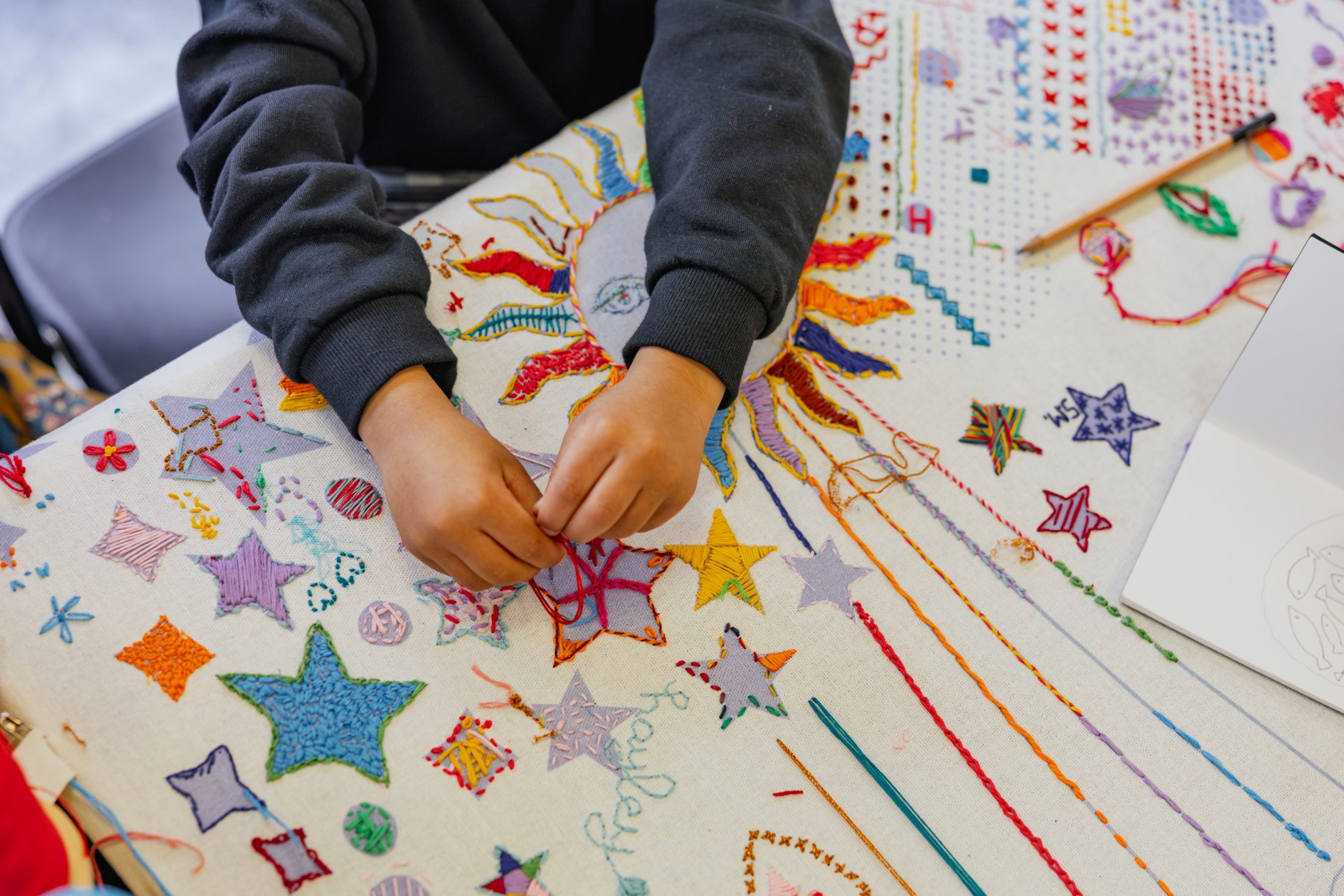 A person is hand-sewing colorful patterns on fabric, featuring stars and a sun design.