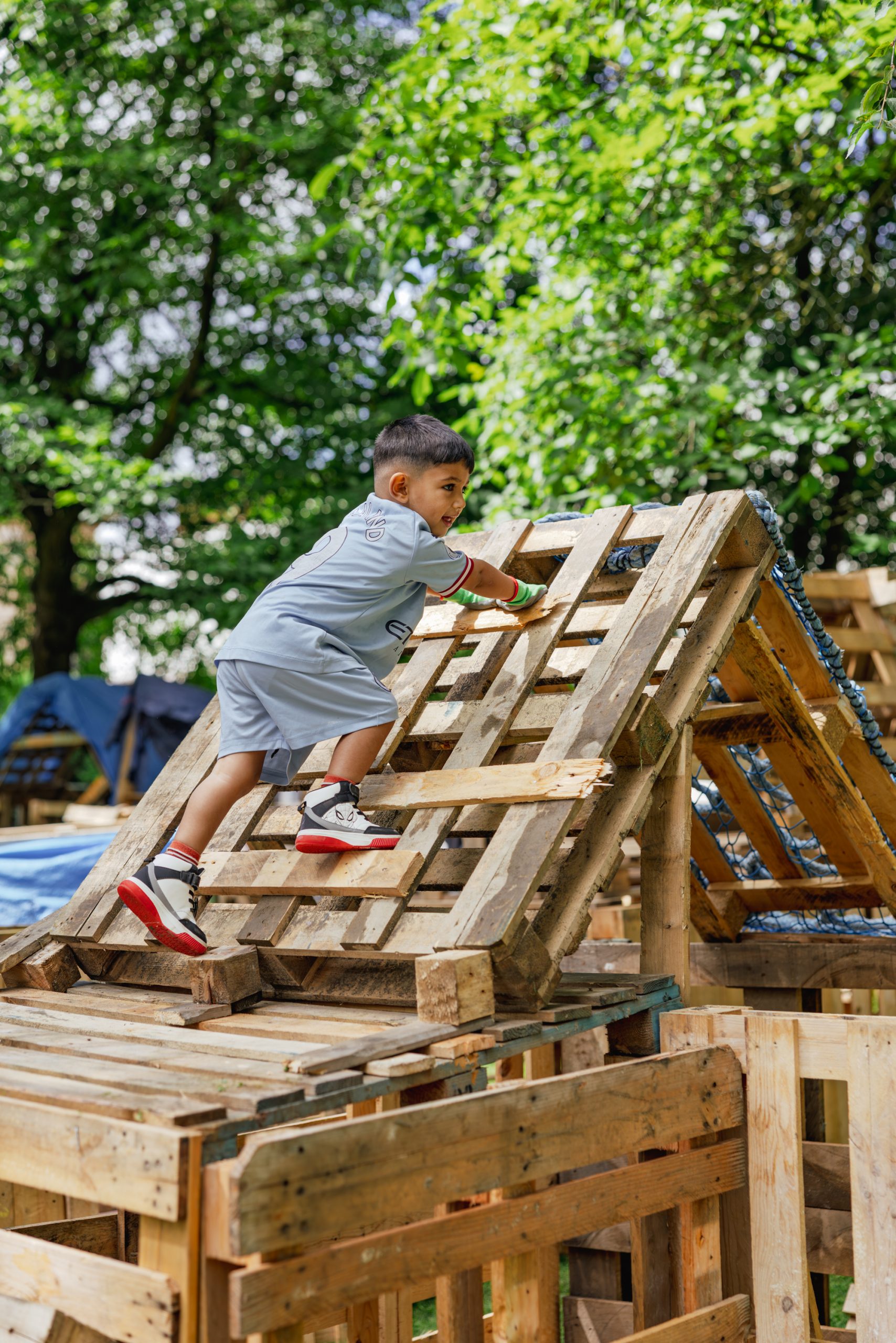 A child climbs a wooden structure, surrounded by green trees.