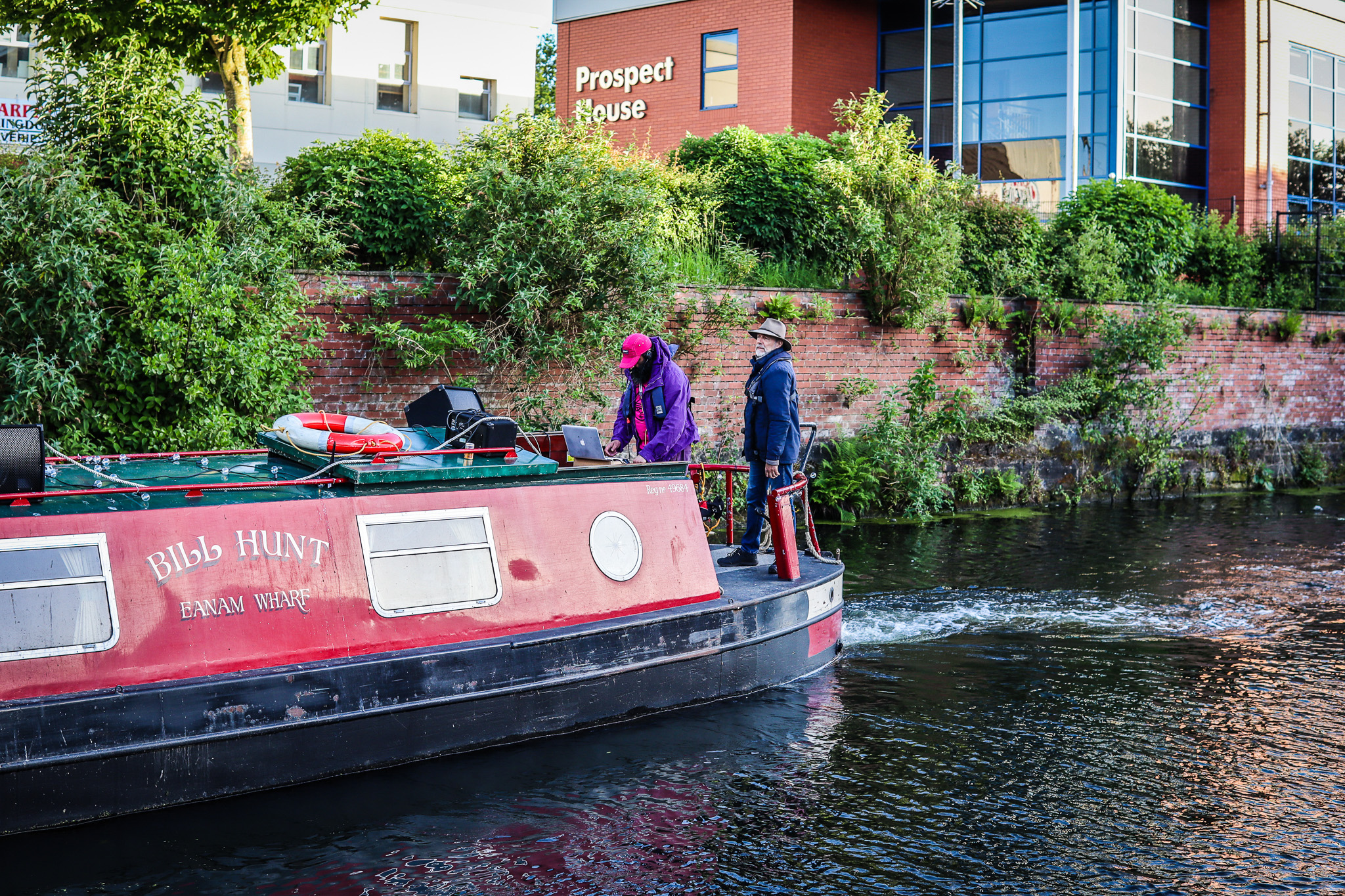 Red canal boat moves along a waterway beside a brick wall and lush greenery, with two people on deck, one DJing.