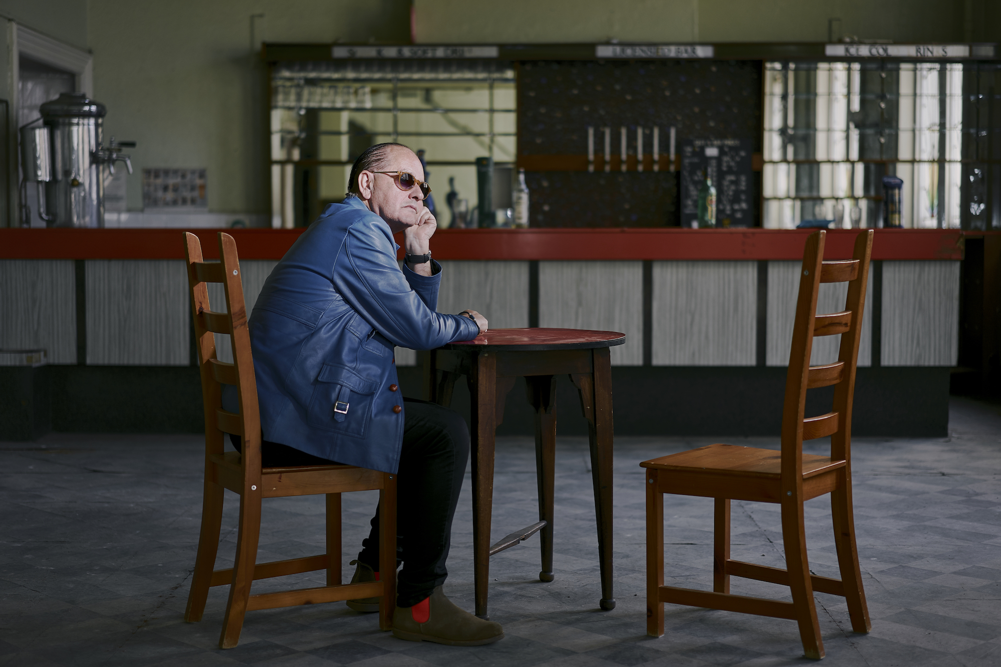 Martyn Ware in a blue jacket and sunglasses sits at a wooden table in an empty retro bar with a red counter and mirrored backdrop.