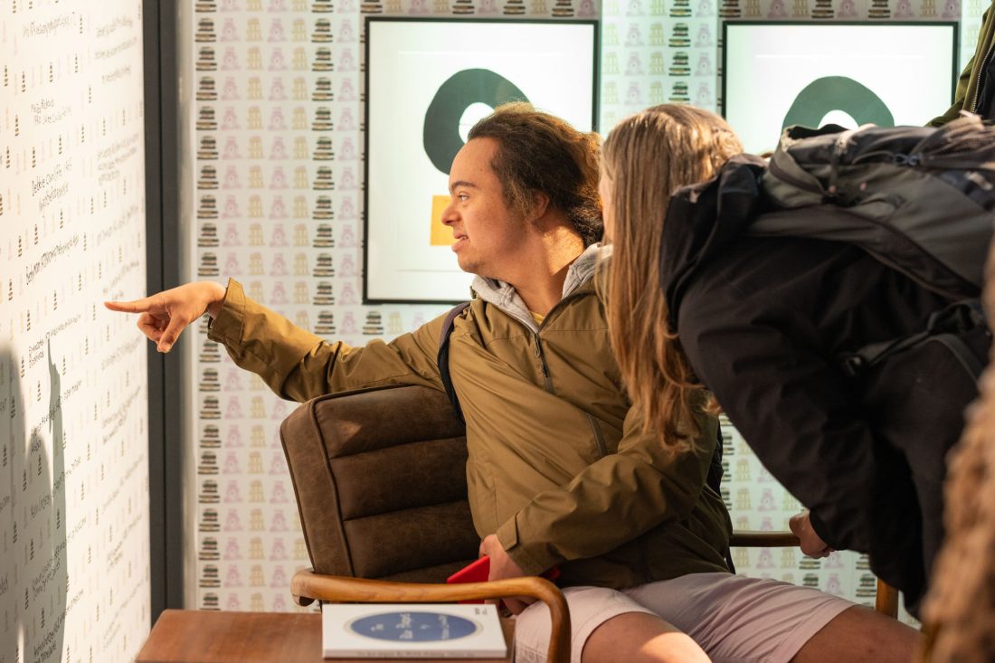 A person sitting and pointing at a wall whilst another person with a rucksack leans in to look. Both appear focused, and there are framed artworks hanging behind them.