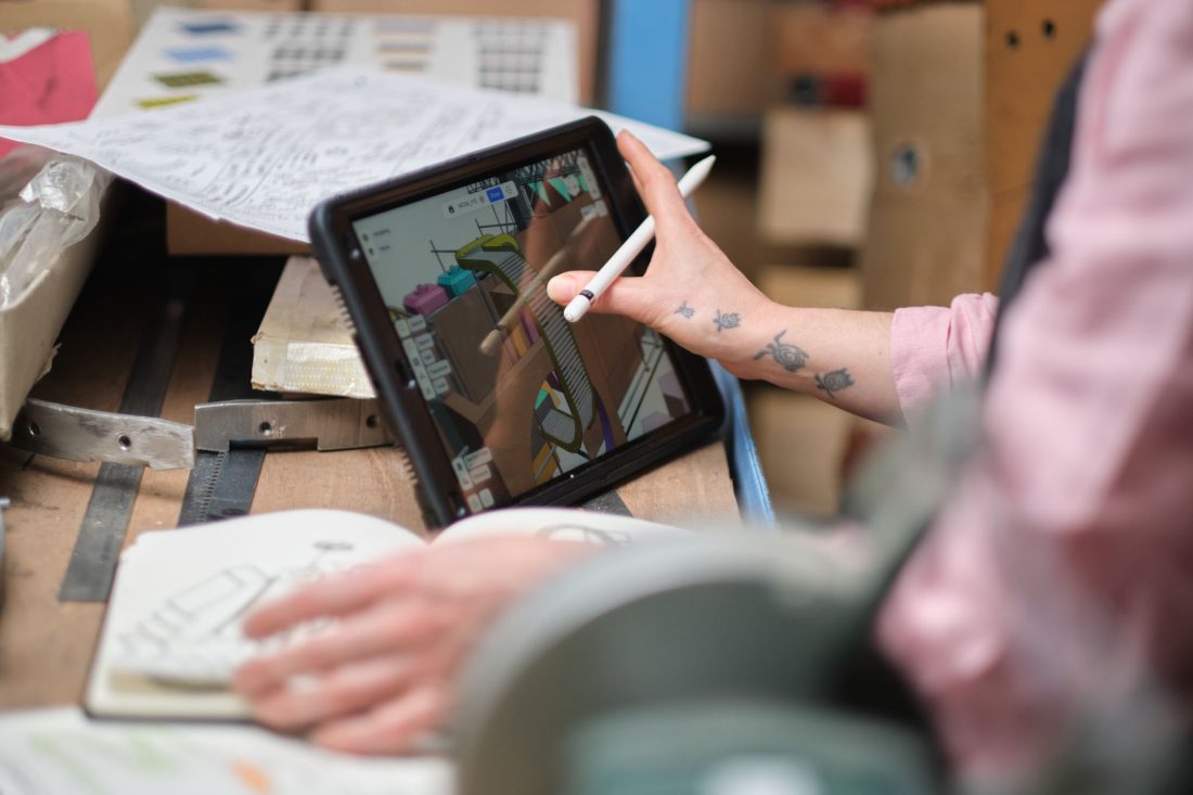 A person with turtle tattoos on their forearm uses a stylus on a tablet displaying 3D design software. The tablet rests on a messy wooden workbench cluttered with paper sketches, a metal tool, and cardboard boxes in the background.