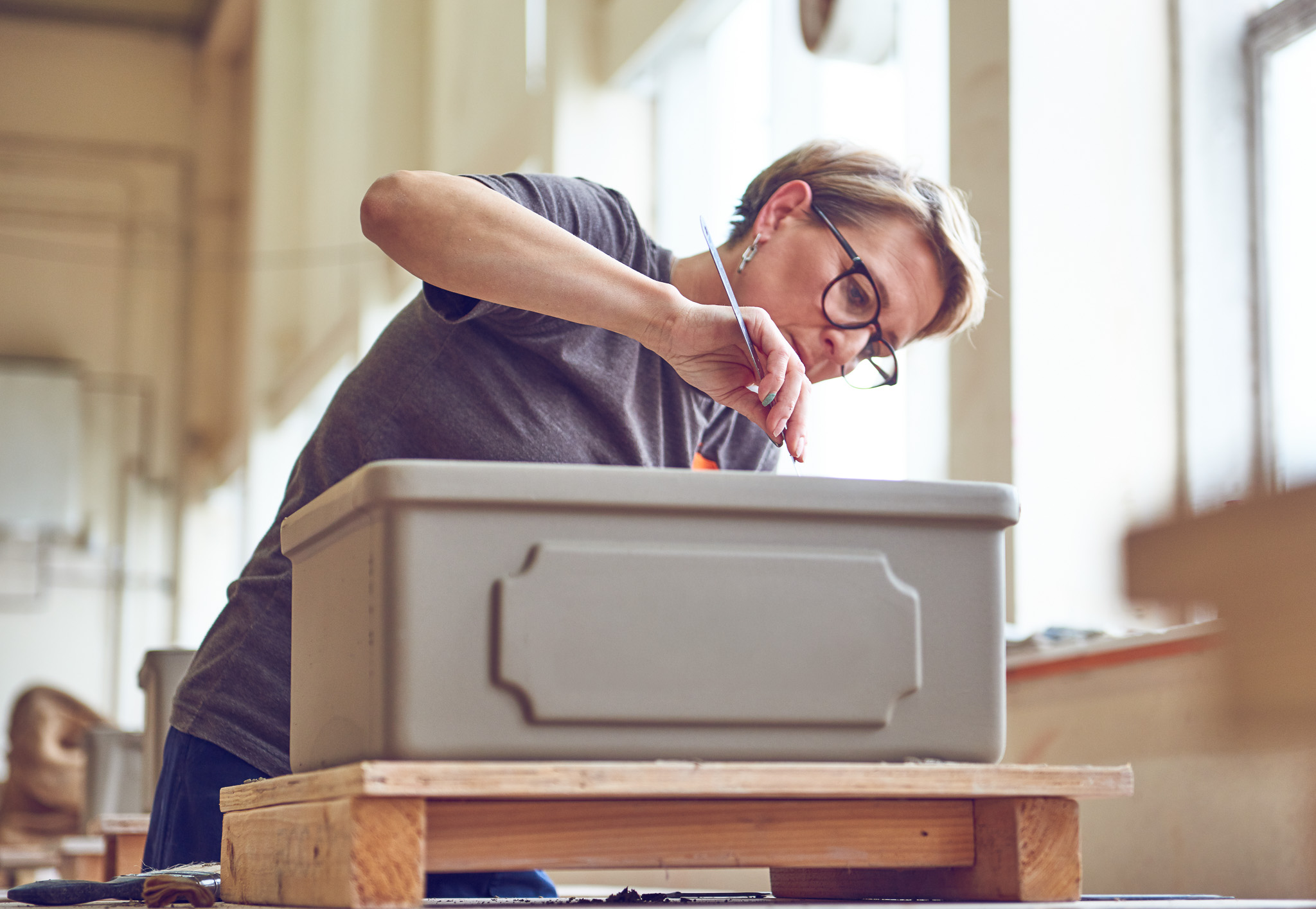 A focused person with glasses works on a clay sink, using a small sculpting tool.