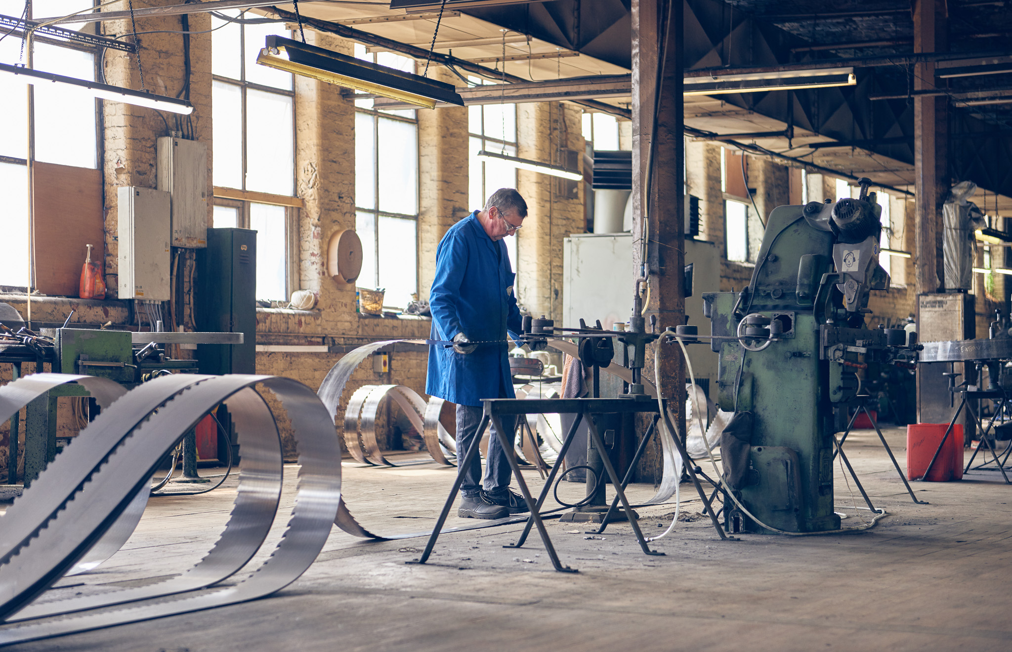 A man in a blue coat works in an industrial workshop, bending metal strips using machinery.