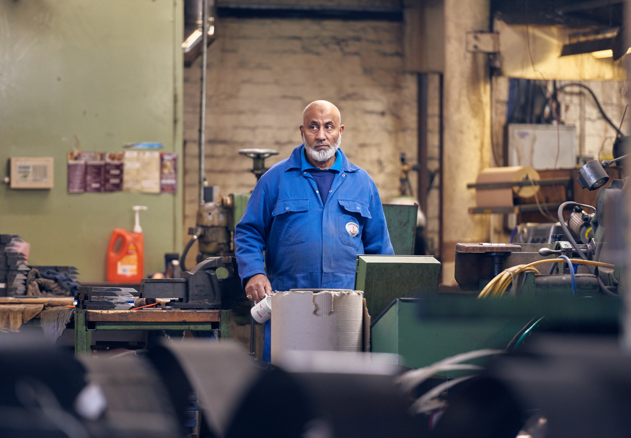 Elderly man in a blue work uniform stands in a cluttered workshop, surrounded by machinery and tools.