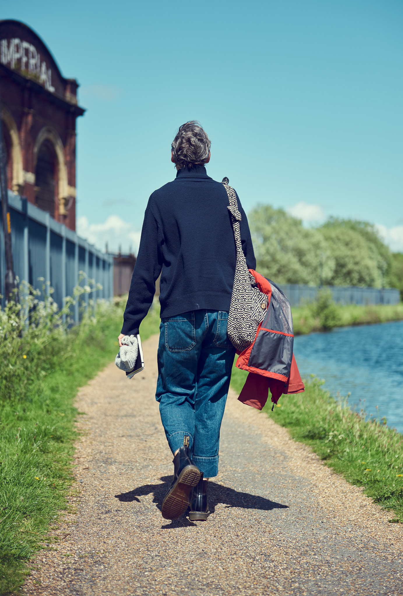 A person walks away on a sunny path by a canal, carrying a red jacket and bag.