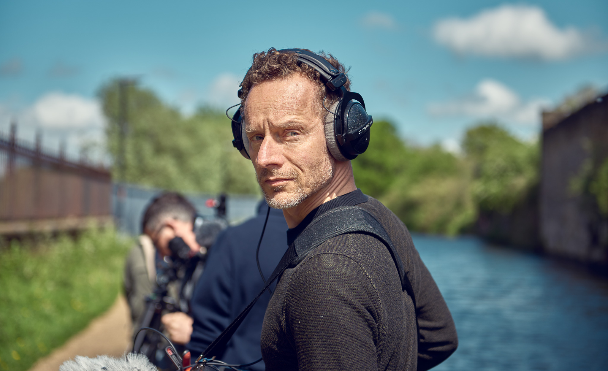 A man wearing headphones stands near a canal, holding audio equipment. Another person in the background operates a camera.
