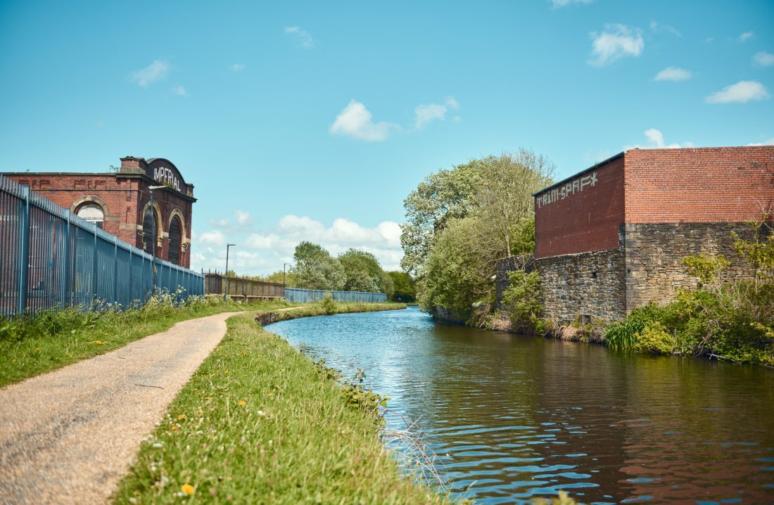 Canal path with grassy verge leading to a distant bridge, flanked by red brick buildings. The sky is blue with scattered clouds.