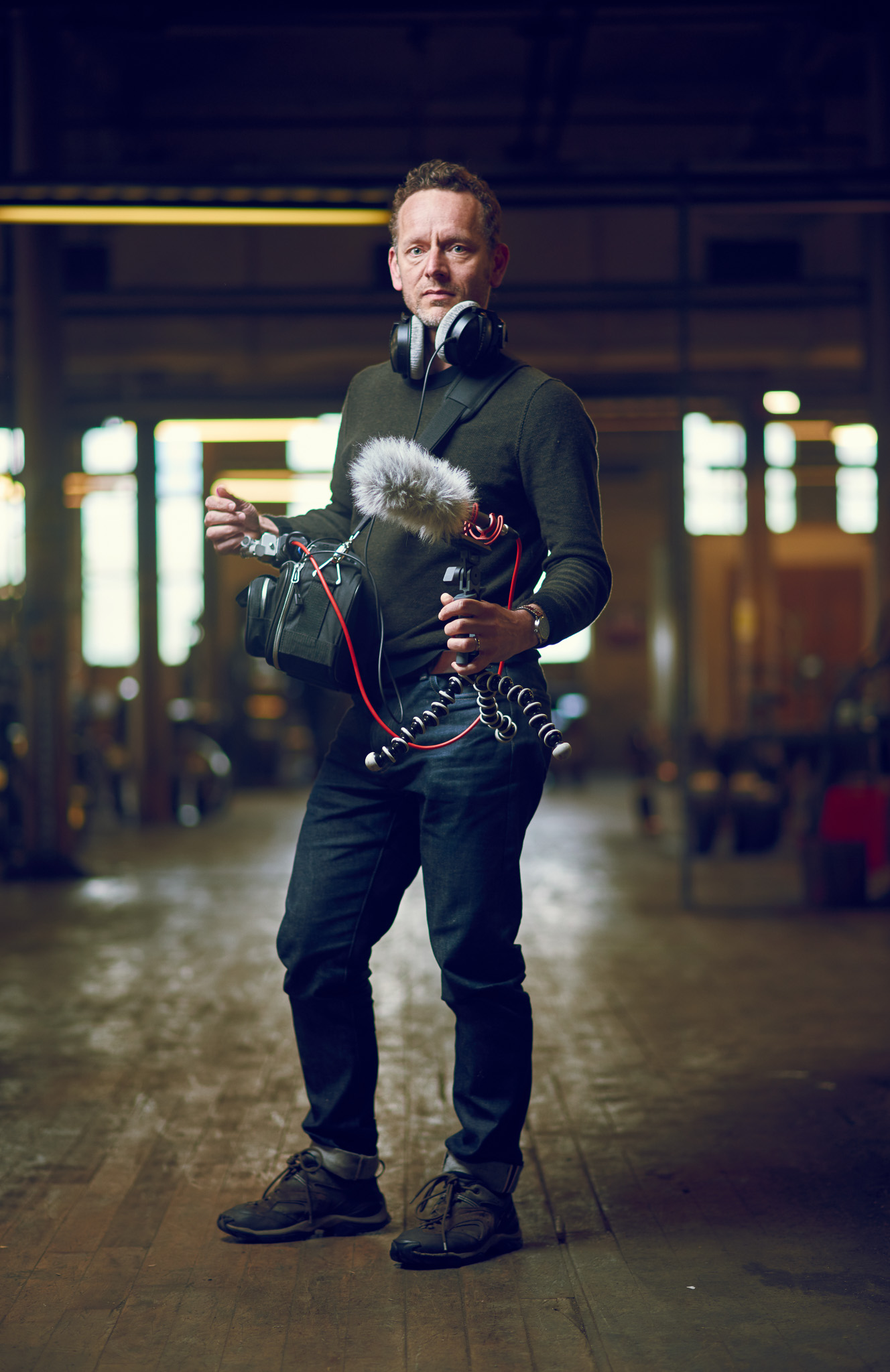 A person stands in a dimly lit space holding audio recording equipment, wearing headphones and a microphone.
