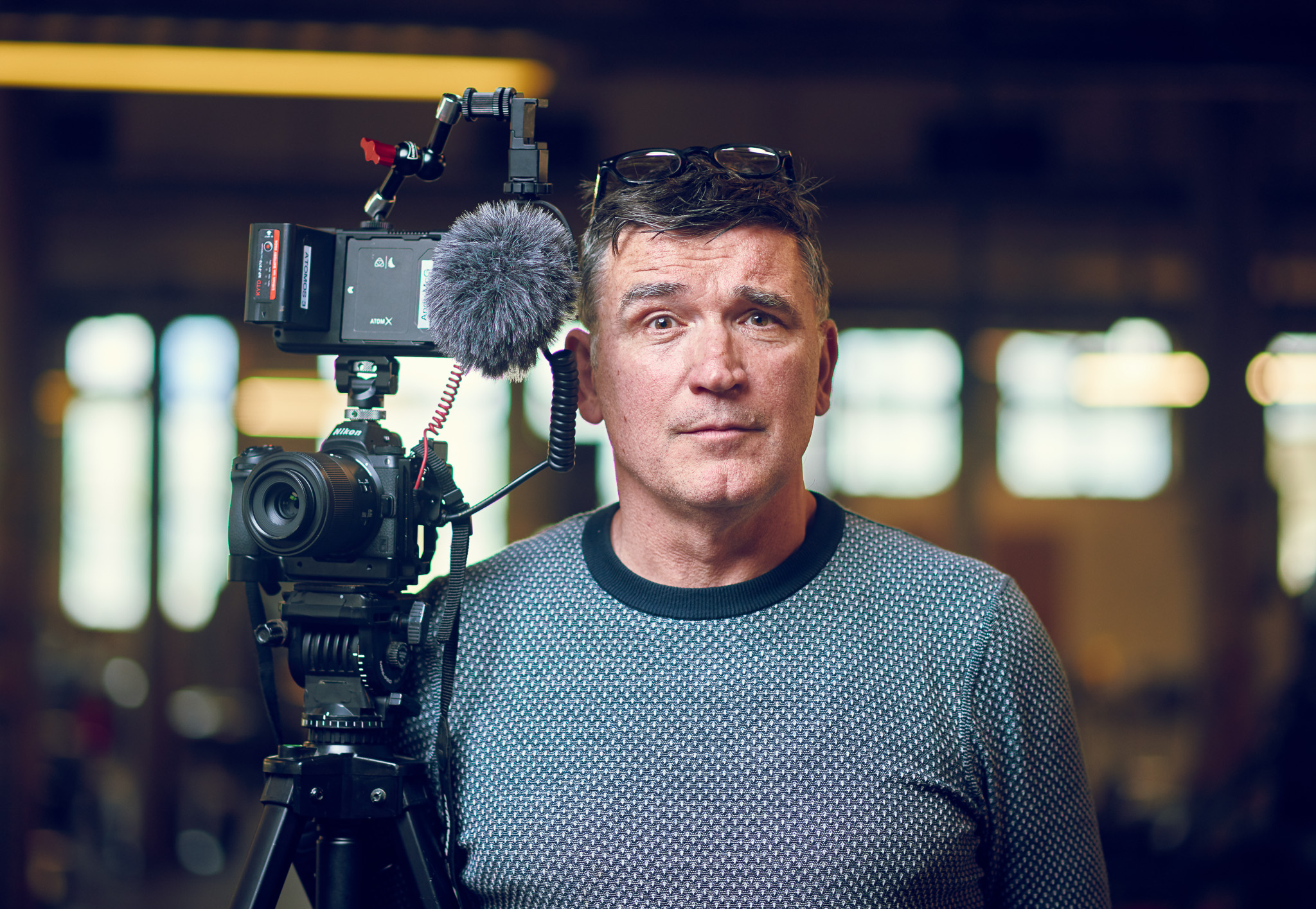 A man stands indoors beside a professional video camera with a microphone. He wears a patterned sweater, looking focused. The background is softly blurred.