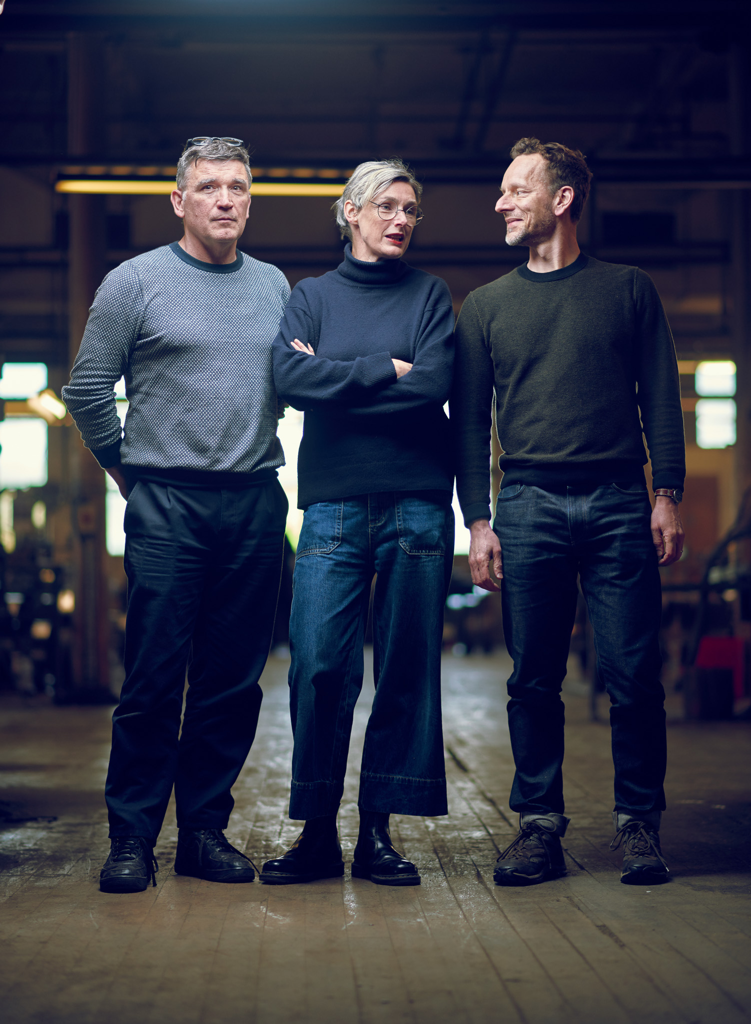 Three people stand indoors in casual clothing. They appear relaxed, with two smiling at each other.