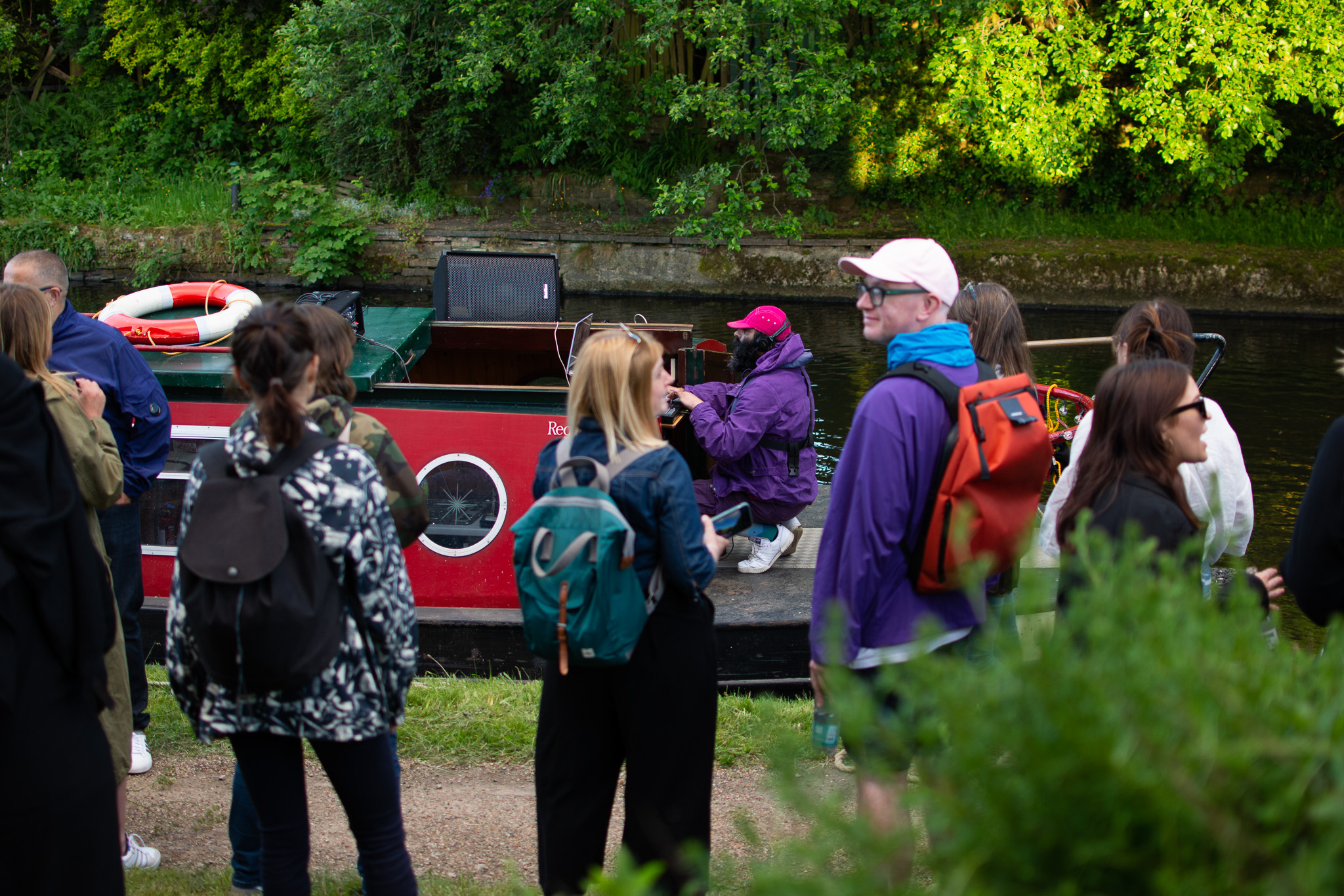 A group of people stand near a red canal boat on a sunny day. The boat is moored alongside a green, wooded bank. One person sits on the boat playing music.