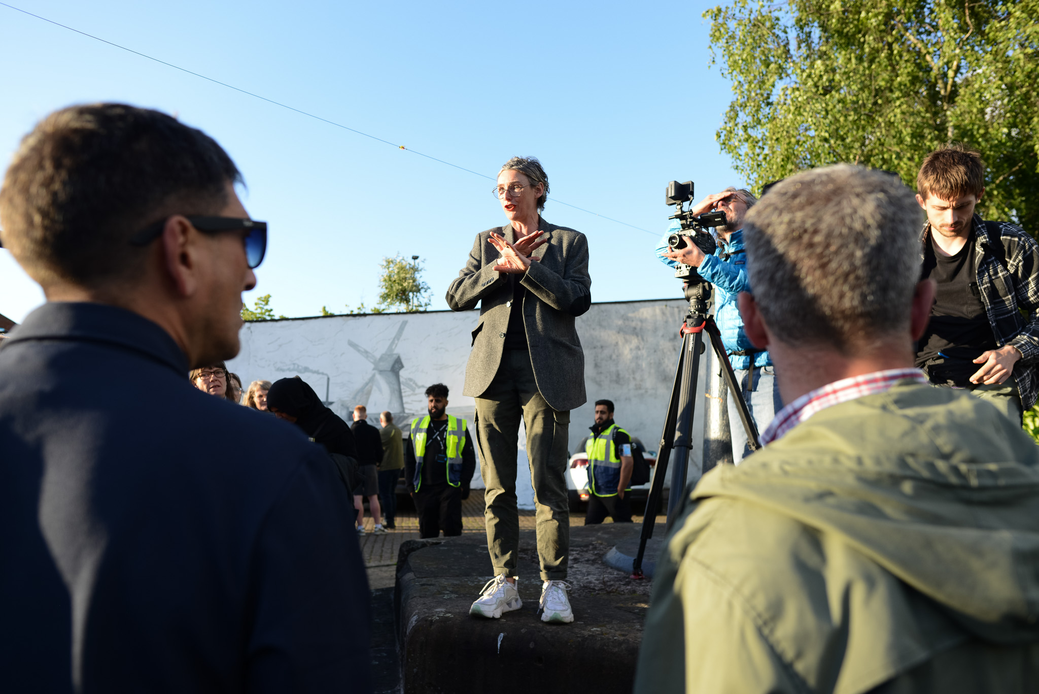 Jacqueline Donachie stands on a pedestal, gesturing enthusiastically. A film crew is nearby, and there are spectators and trees in the background.
