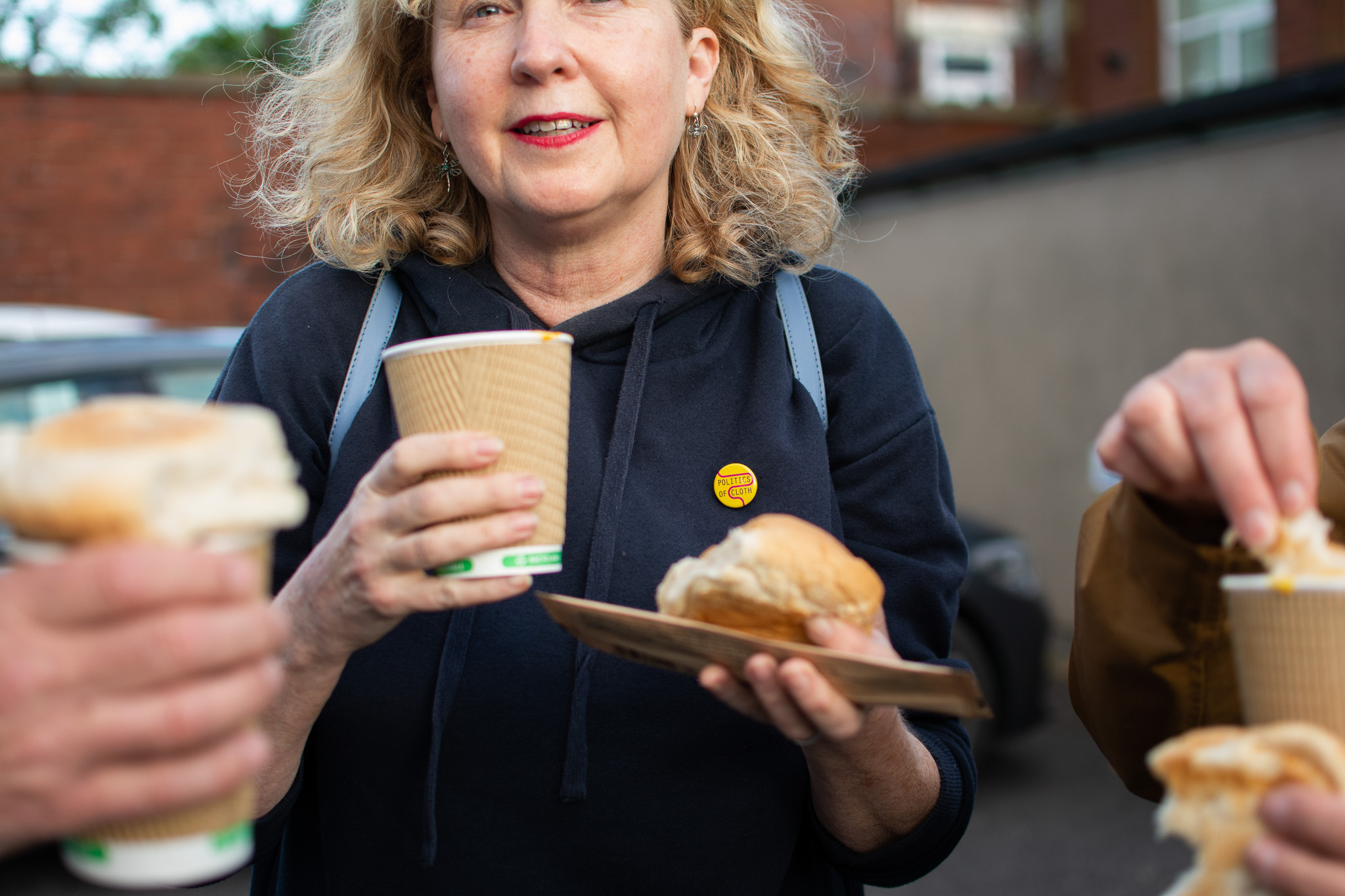 Woman with curly hair holding a paper cup and a bread roll outdoors.