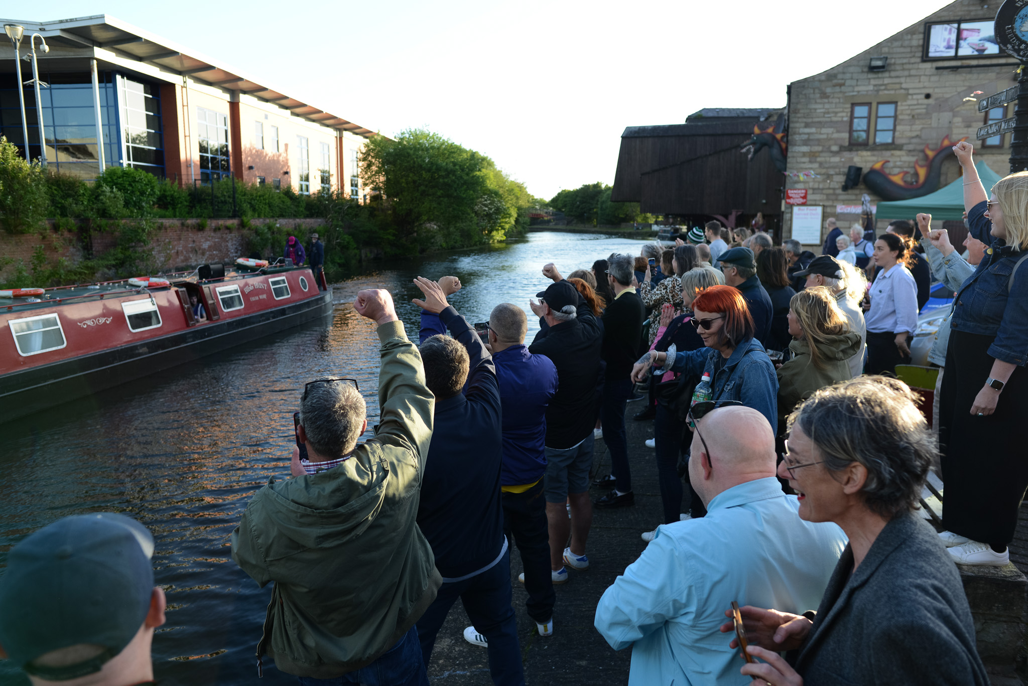 A group of people excitedly wave at a passing canal boat on a sunny day. The crowd stands on the canal bank, exuding a lively and cheerful atmosphere.