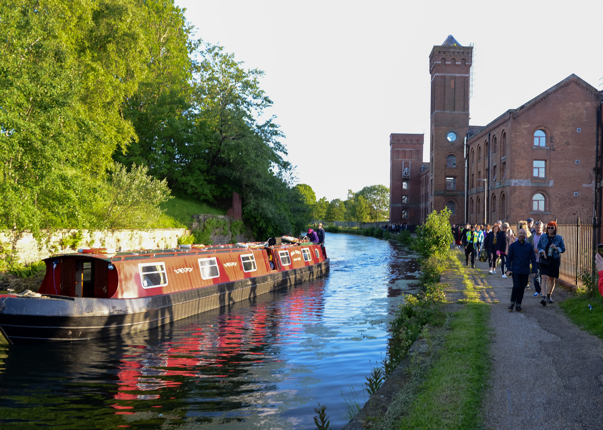 A red narrowboat floats on a serene canal beside a historic brick building. People walk along the towpath, surrounded by lush greenery in sunlight.