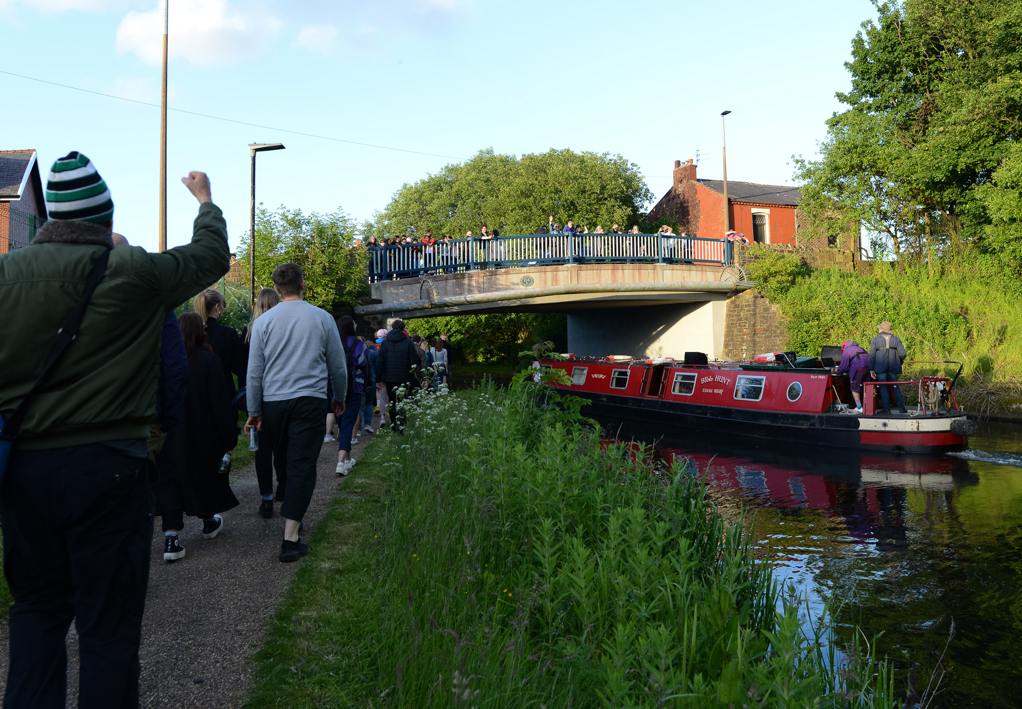 A lively canal scene with people walking along a path, some waving. A red narrowboat passes under a bridge, surrounded by greenery and a sunny sky.
