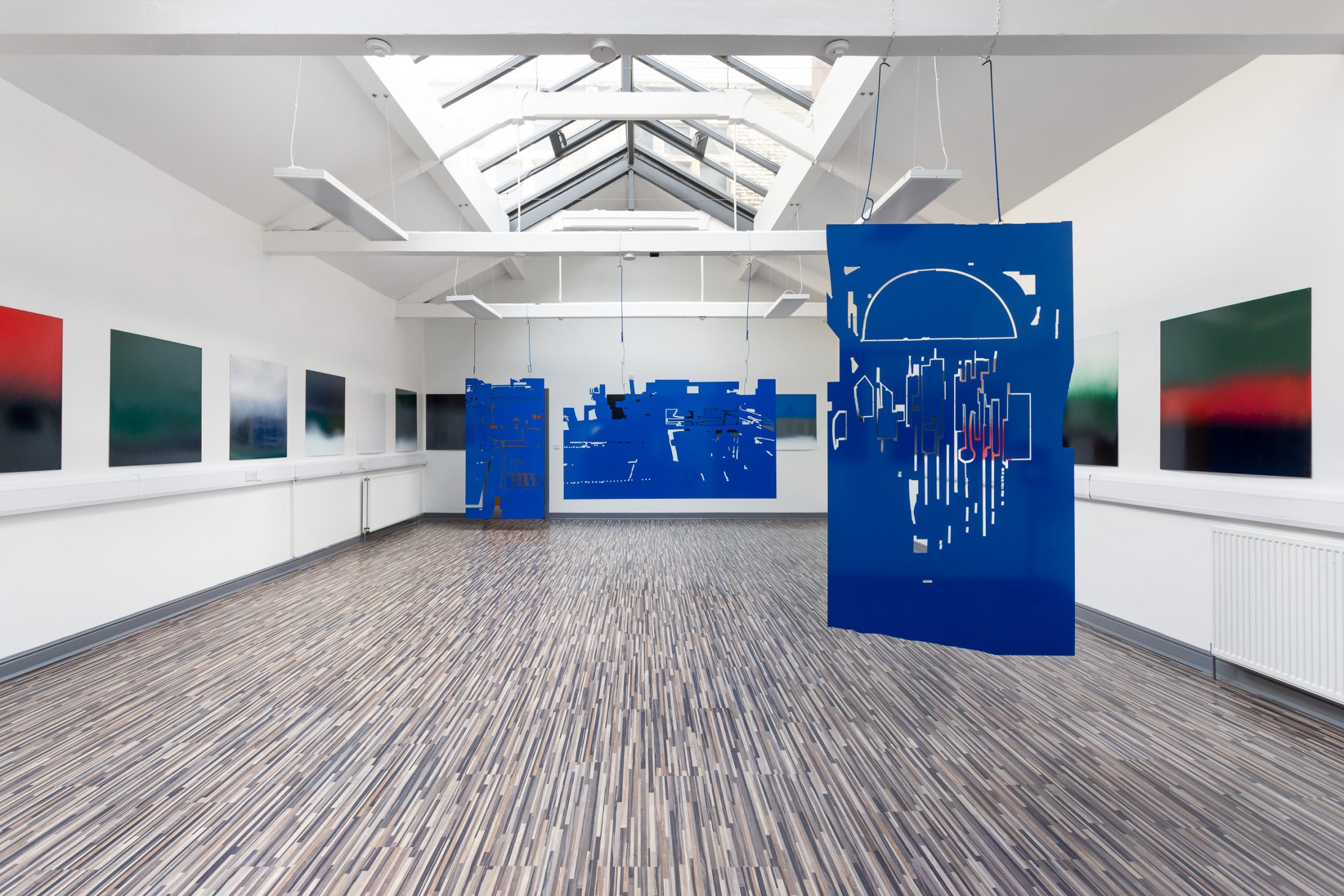 Suspended blue metal panels with cut-out patterns hang in a large gallery space, surrounded by abstract gradient paintings on the walls.
