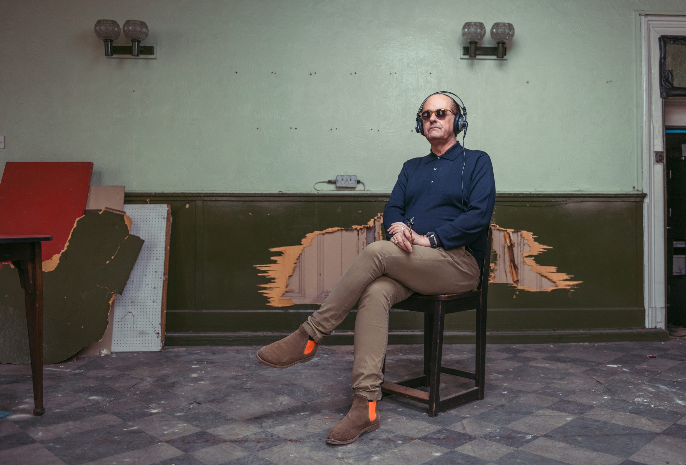 Martyn Ware in sunglasses and headphones sits cross-legged on a chair in a worn room with peeling paint.