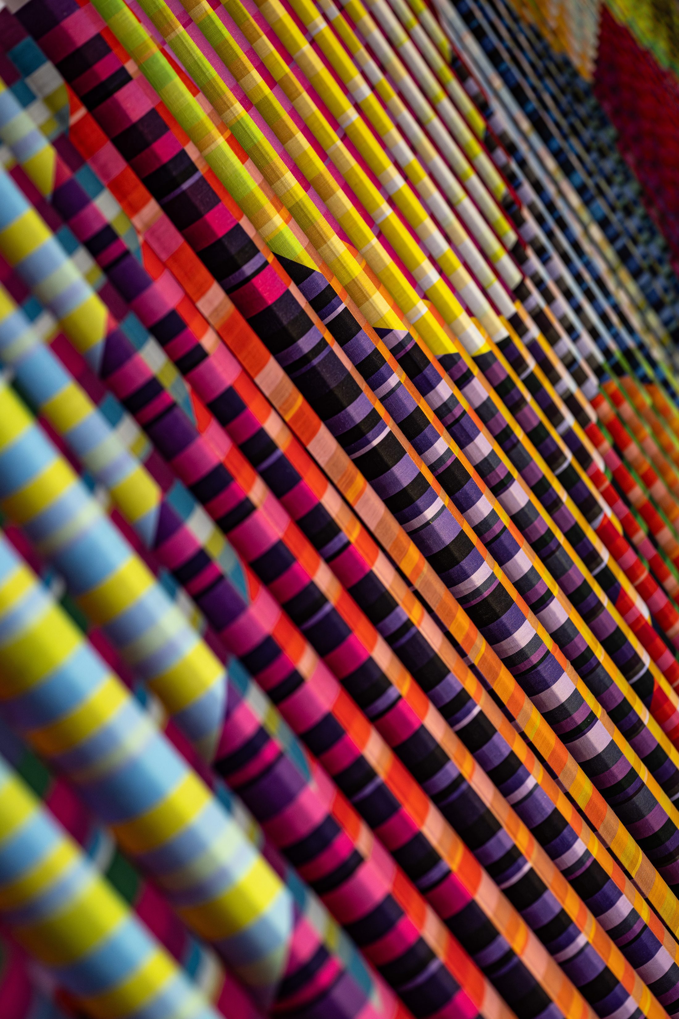 Colourful abstract artwork featuring diagonal rows of vibrant, striped tubes in yellow, red, blue, purple, and pink, creating a dynamic, layered pattern with a sense of movement and depth.