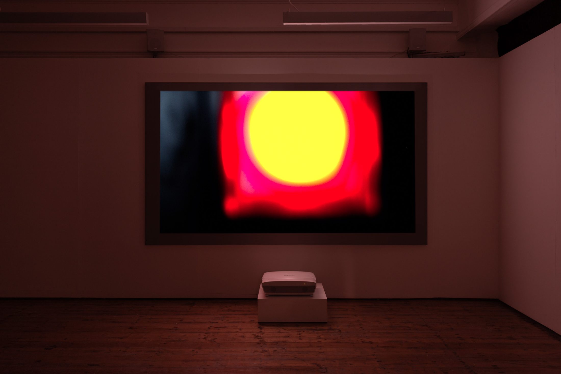 Large screen projects a vivid abstract image with a bright yellow circle surrounded by red and black hues in a dimly lit gallery.