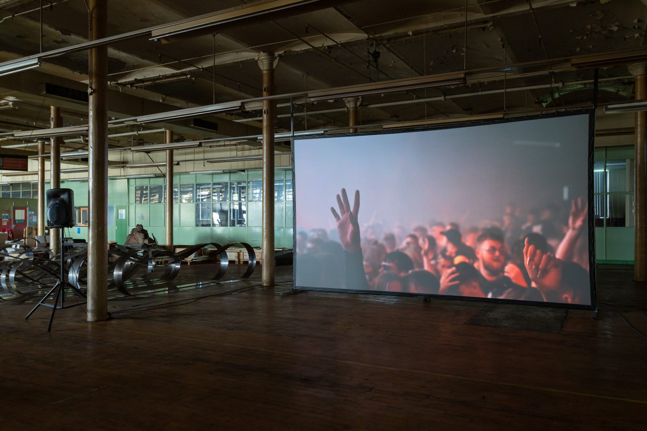 Large screen displays a crowd with raised hands in a dimly lit industrial space, with metal coils and a speaker visible.