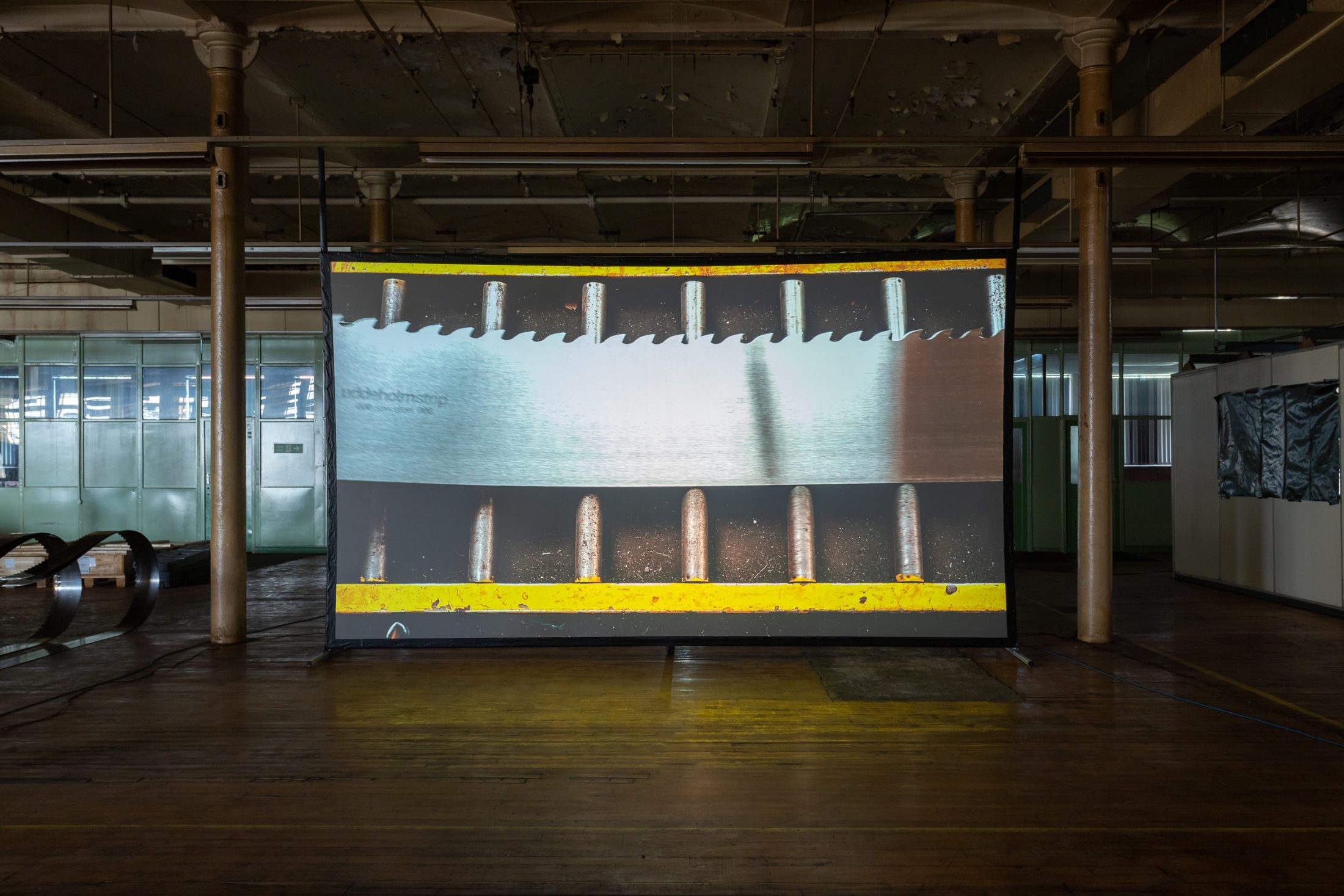 Large projection of a close-up saw blade displayed in an industrial space with wooden floors and metal pillars.
