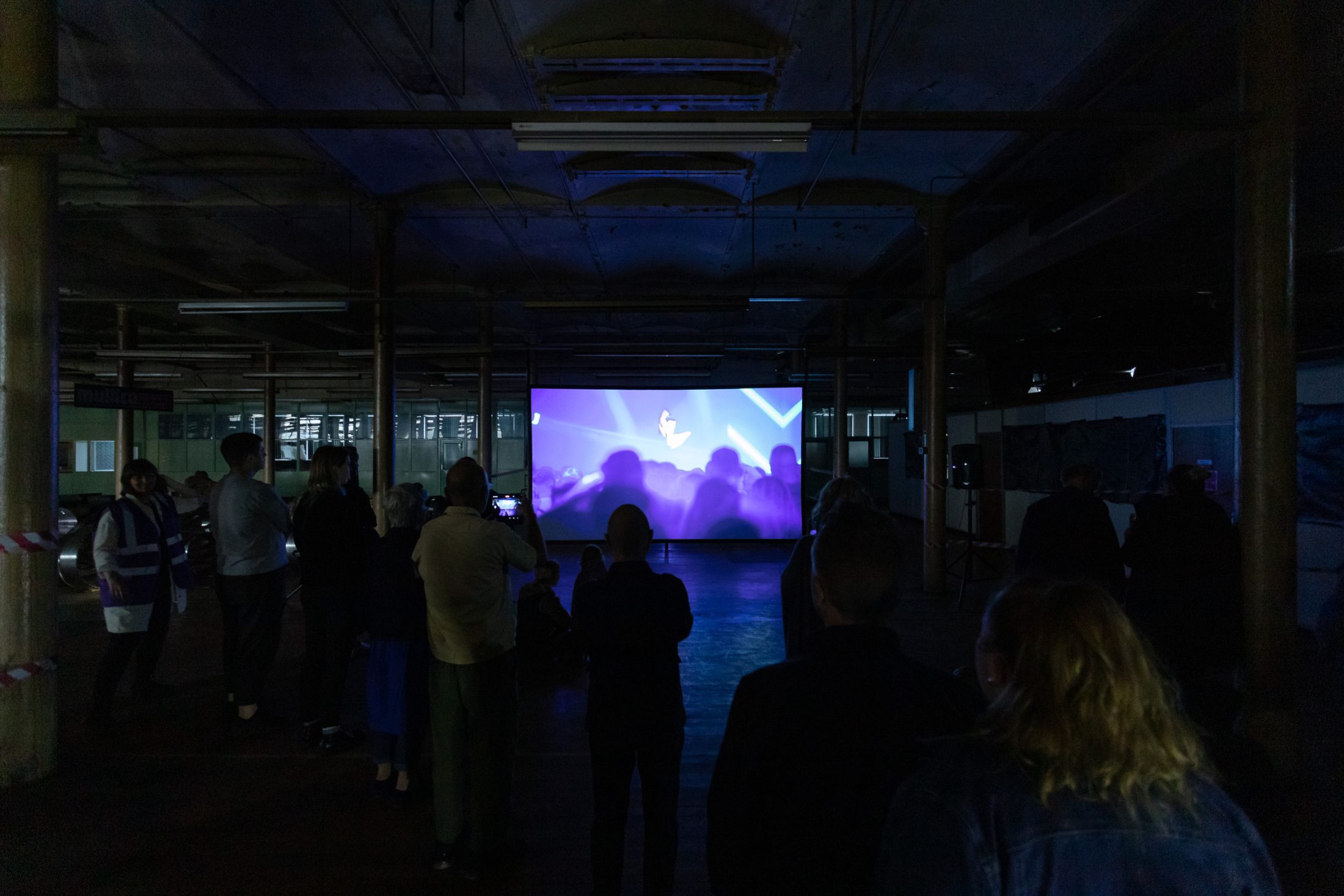 Audience members stand in a dimly lit room, watching a vibrant blue and purple screen projection.