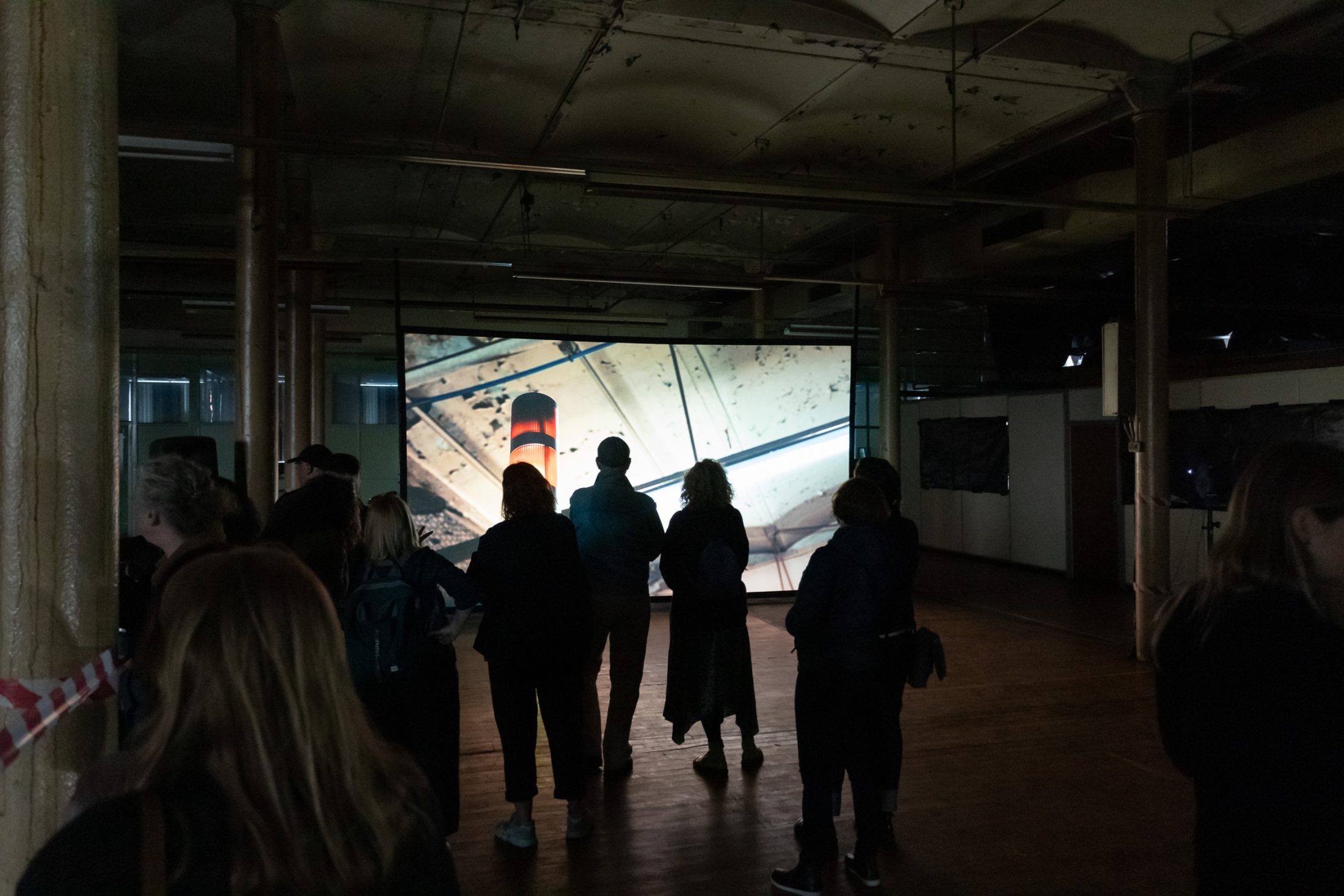 A group of people stand in a dimly lit room, watching a large screen.