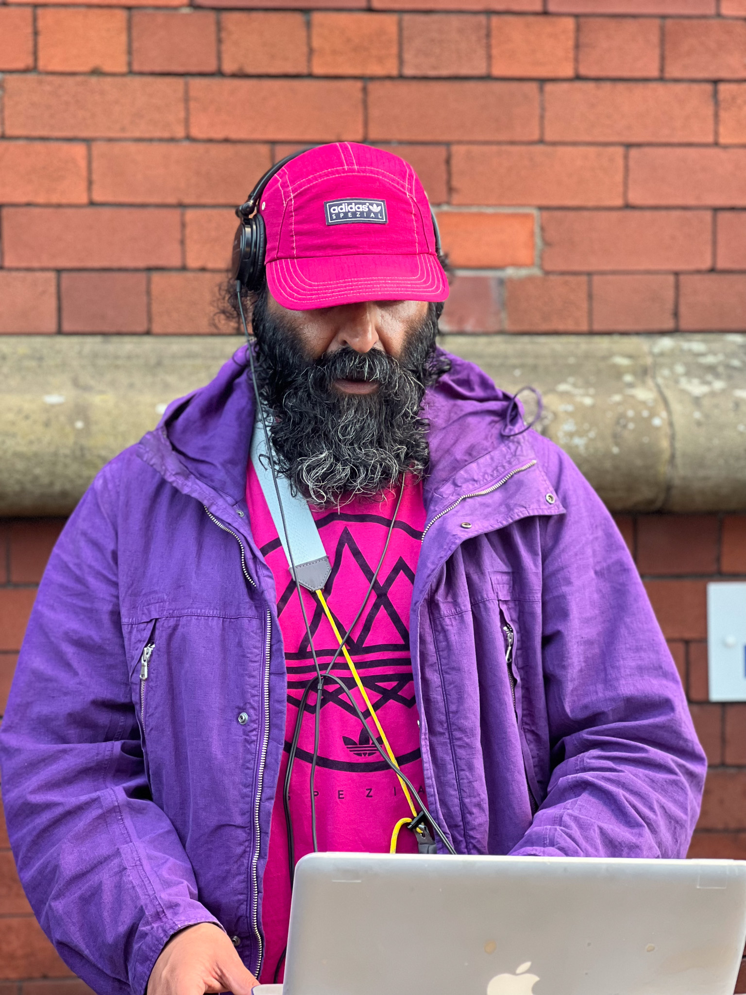 Man wearing a red cap and purple jacket with headphones, standing against a red brick wall, DJing on a laptop.