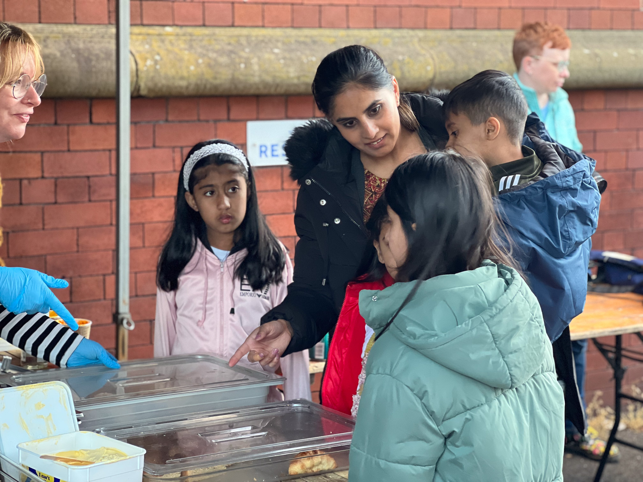 A woman with children selects food at an outdoor stall with covered trays.
