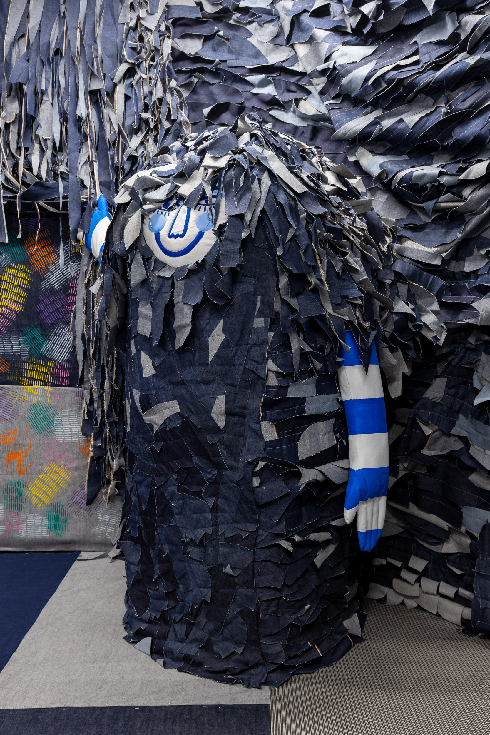 A large, sculpture made of layered, frayed denim strips covers a figure. One hand, covered by a blue and white striped glove, is raised. The background has more hanging denim strips and a colourful, woven pattern on the left.