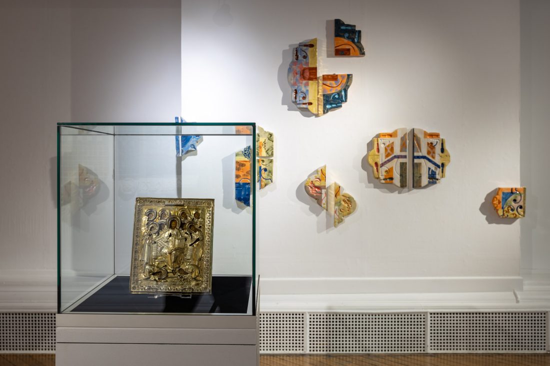 A museum exhibit features a framed religious icon in a glass case and colourful abstract ceramic wall sculptures displayed on a white wall in the background.
