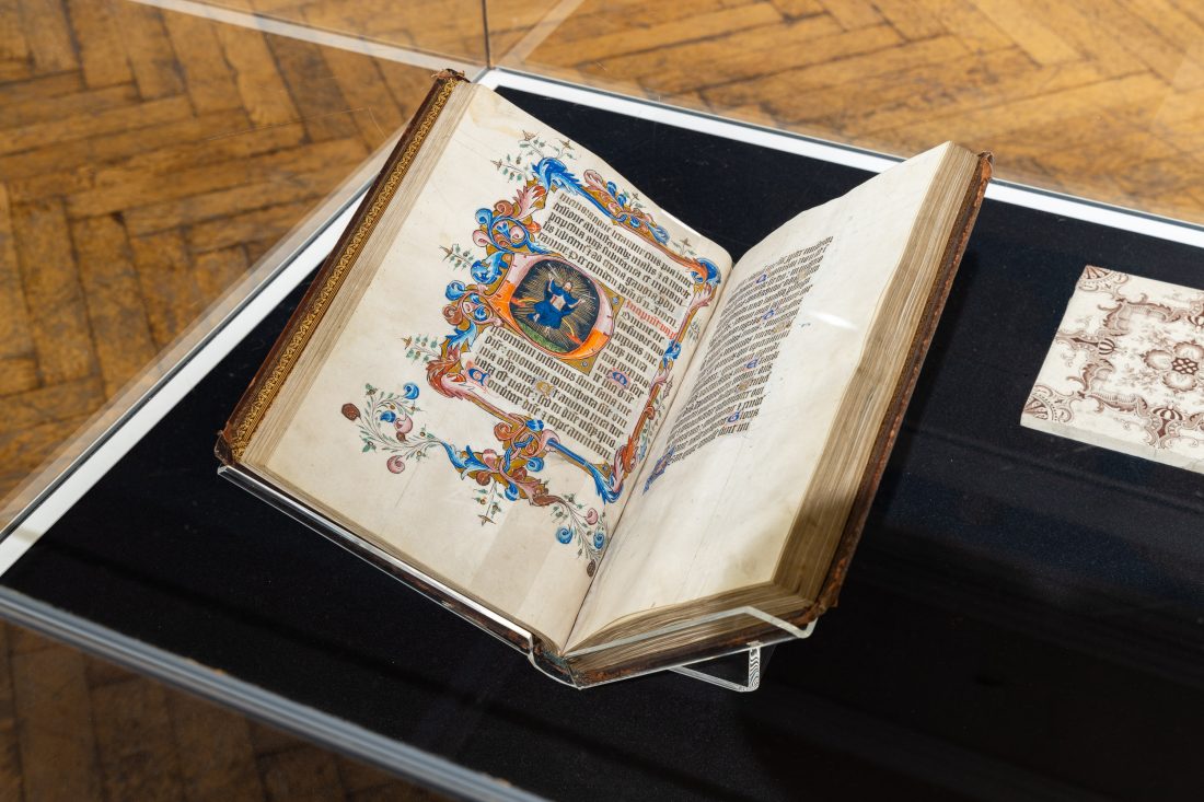 An illuminated manuscript with ornate decorations and a colourful illustration is displayed open in a glass case on a wooden floor.